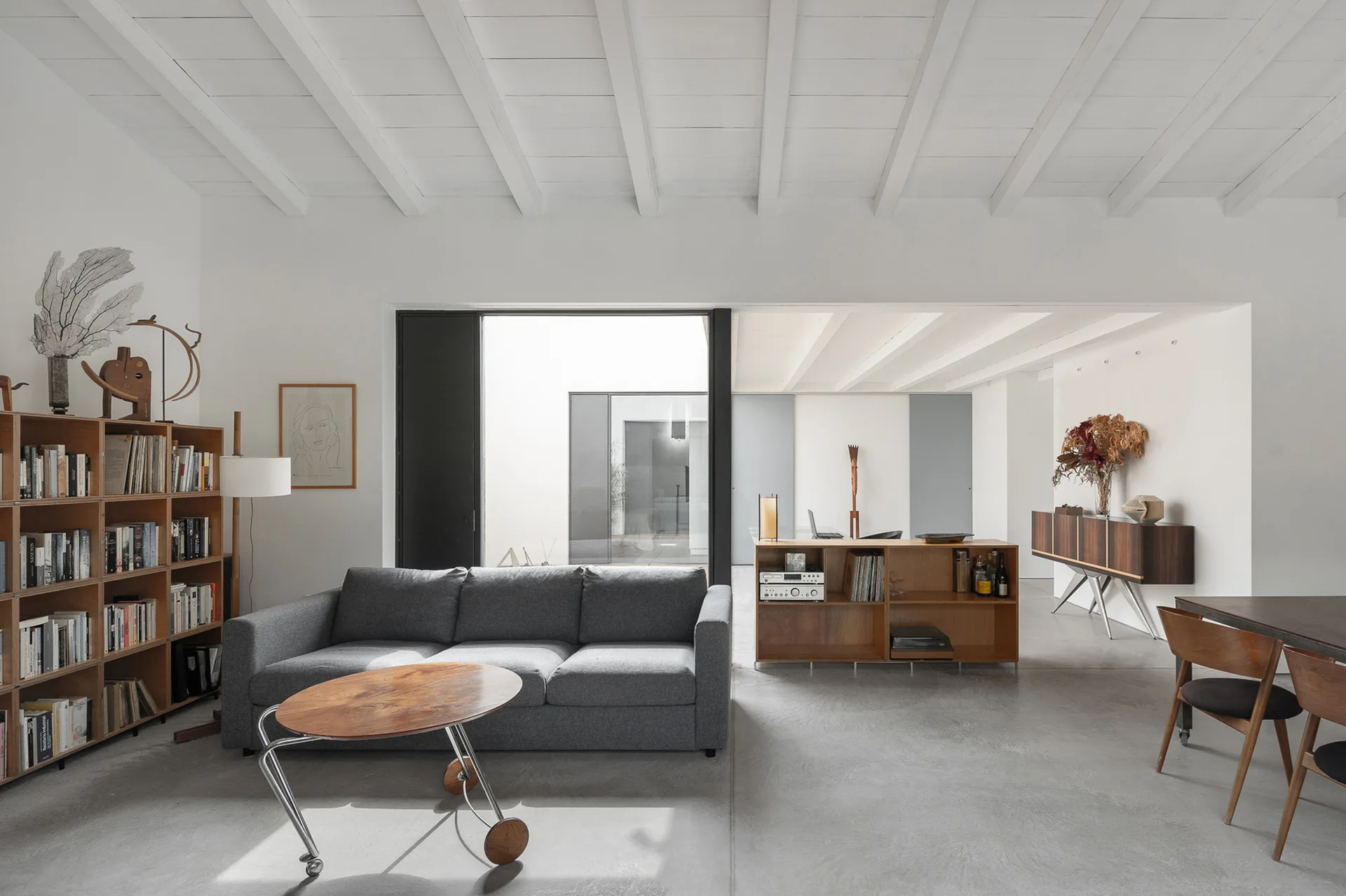 A minimalist living room with a grey fabric sofa, wooden bookshelves, and concrete flooring under white exposed ceiling beams.