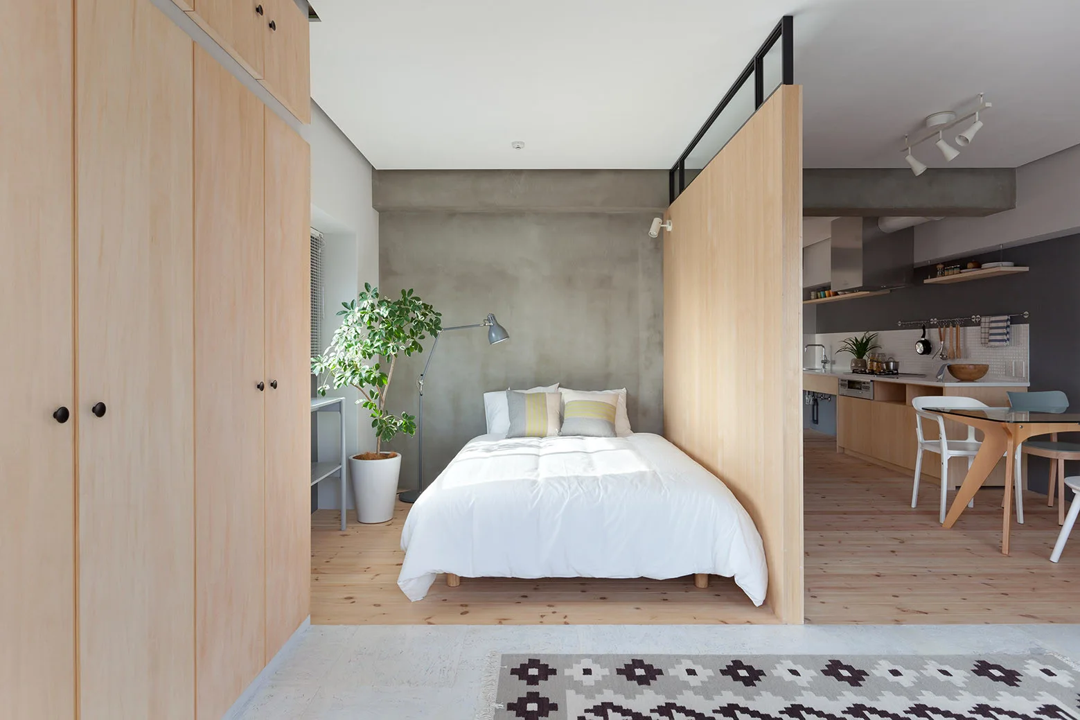A minimalist bedroom in a studio apartment features a plywood partition wall, a bed with white linens, and a raw concrete accent wall.