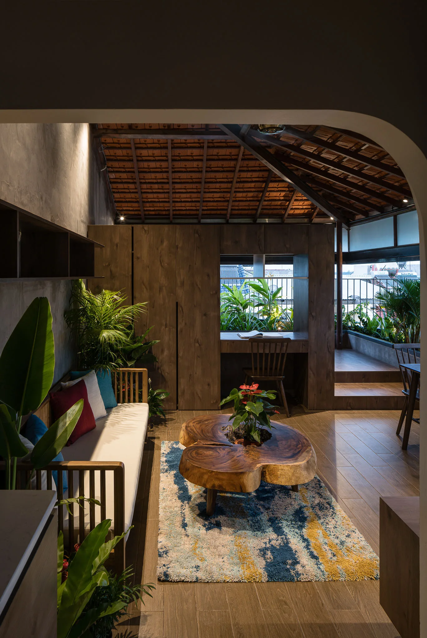 An interior living space with a slatted wood sofa, an organic slab coffee table, and an integrated wooden desk partition looking out to a plant-filled terrace.