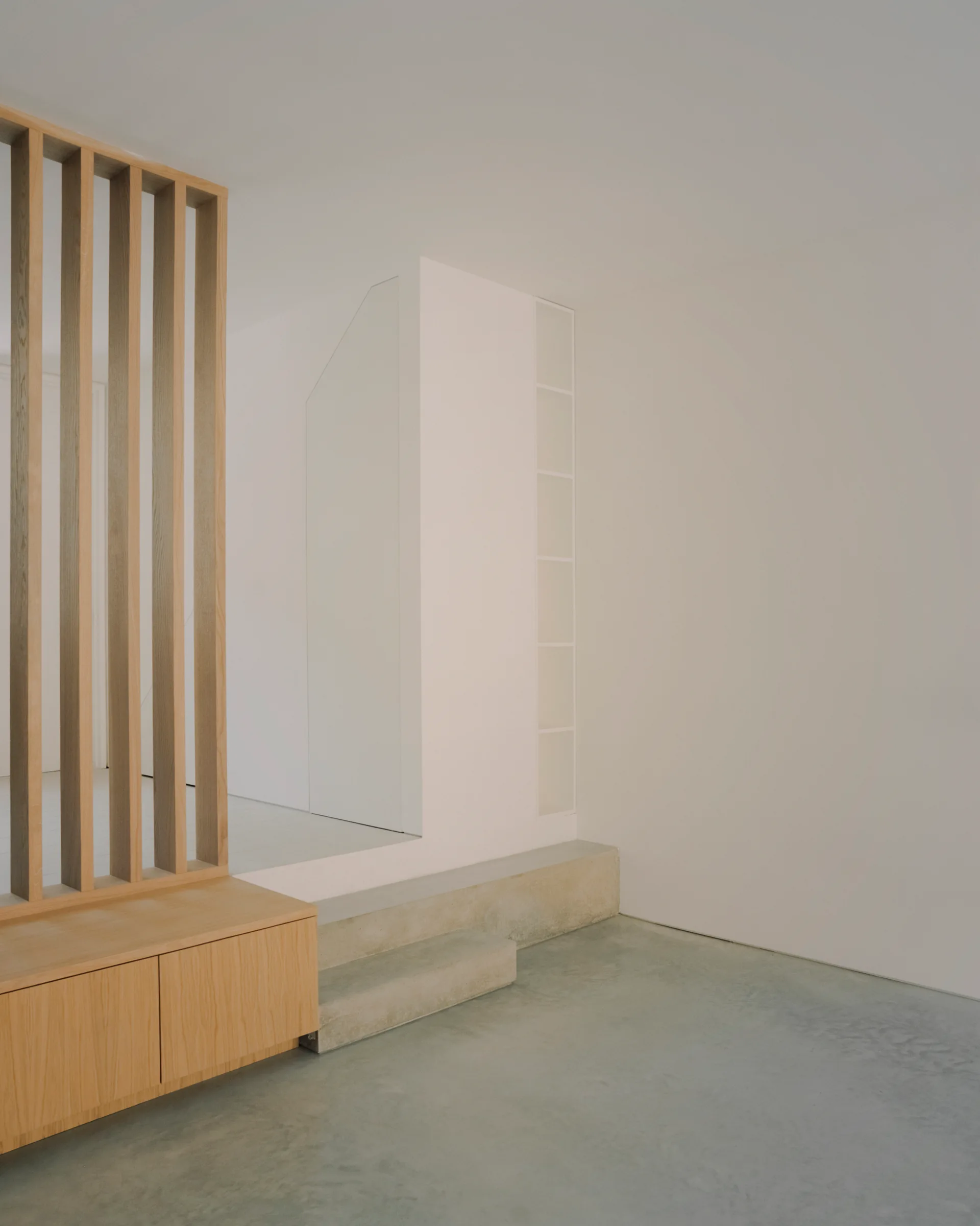 A light wood slatted room divider stands on a low matching wooden cabinet next to smooth grey concrete steps in a minimalist white interior.
