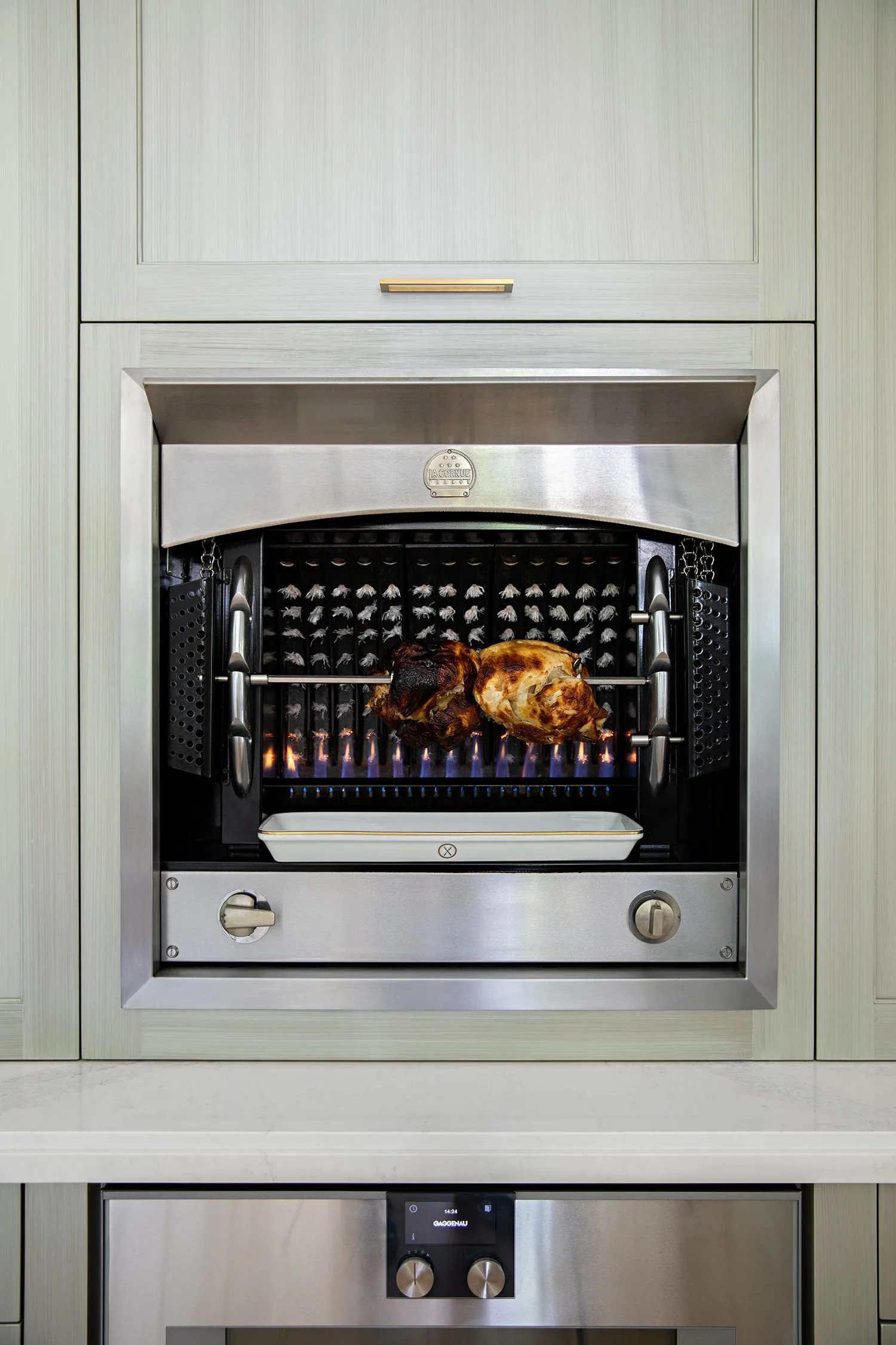 A built-in stainless steel rotisserie oven is roasting chicken inside light grey wood-grain kitchen cabinets above a lower oven panel.