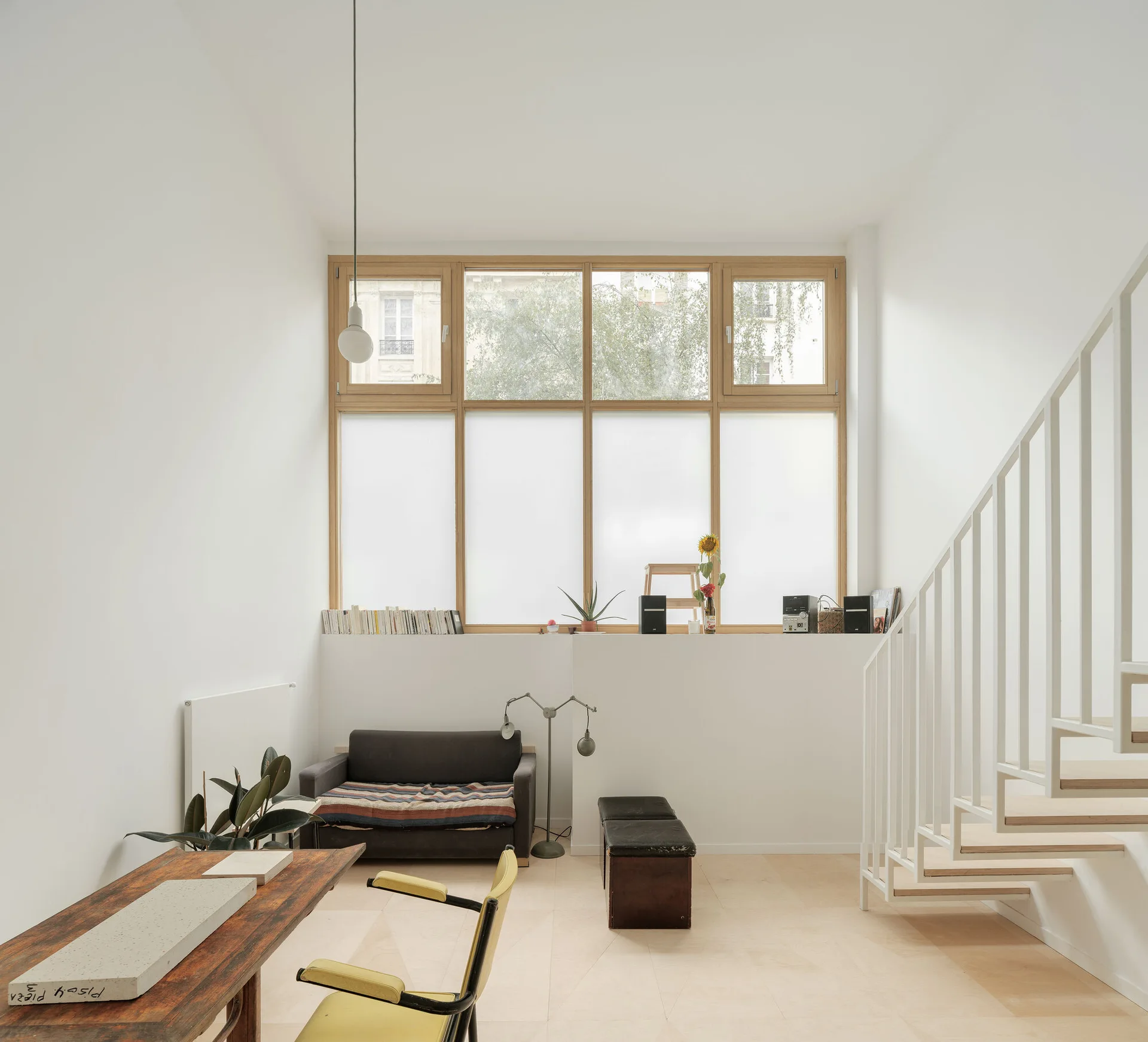A minimalist living room features white walls, a light wood floor, an open-tread staircase, a small dark sofa, and a large wood-framed window.