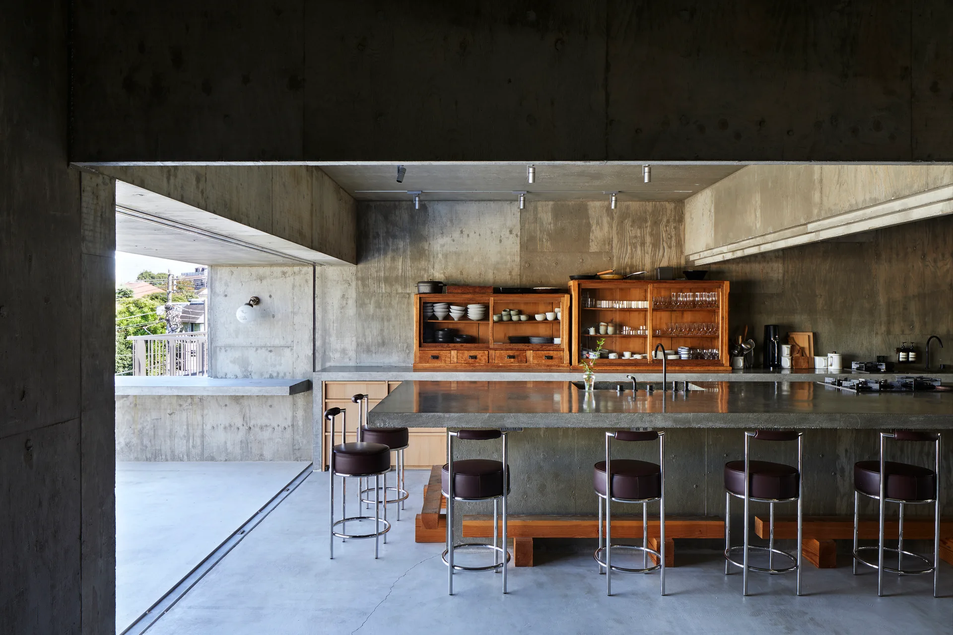 A minimalist kitchen features a long concrete island with leather bar stools set against raw concrete walls and two warm wood storage cabinets.