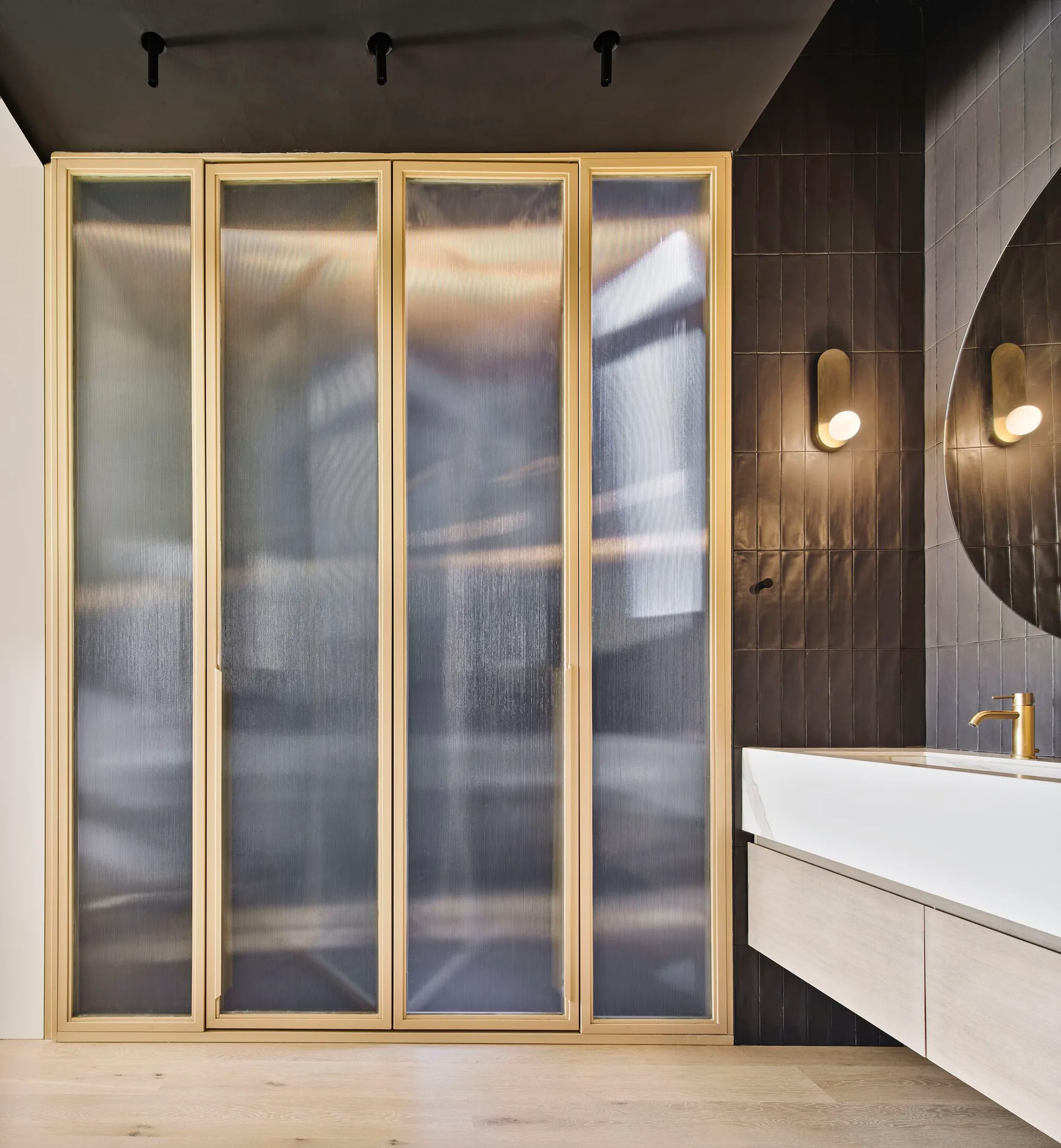 A modern bathroom showing a gold-framed fluted glass partition next to a light wood vanity, dark tiled wall, circular mirror, and gold fixtures.