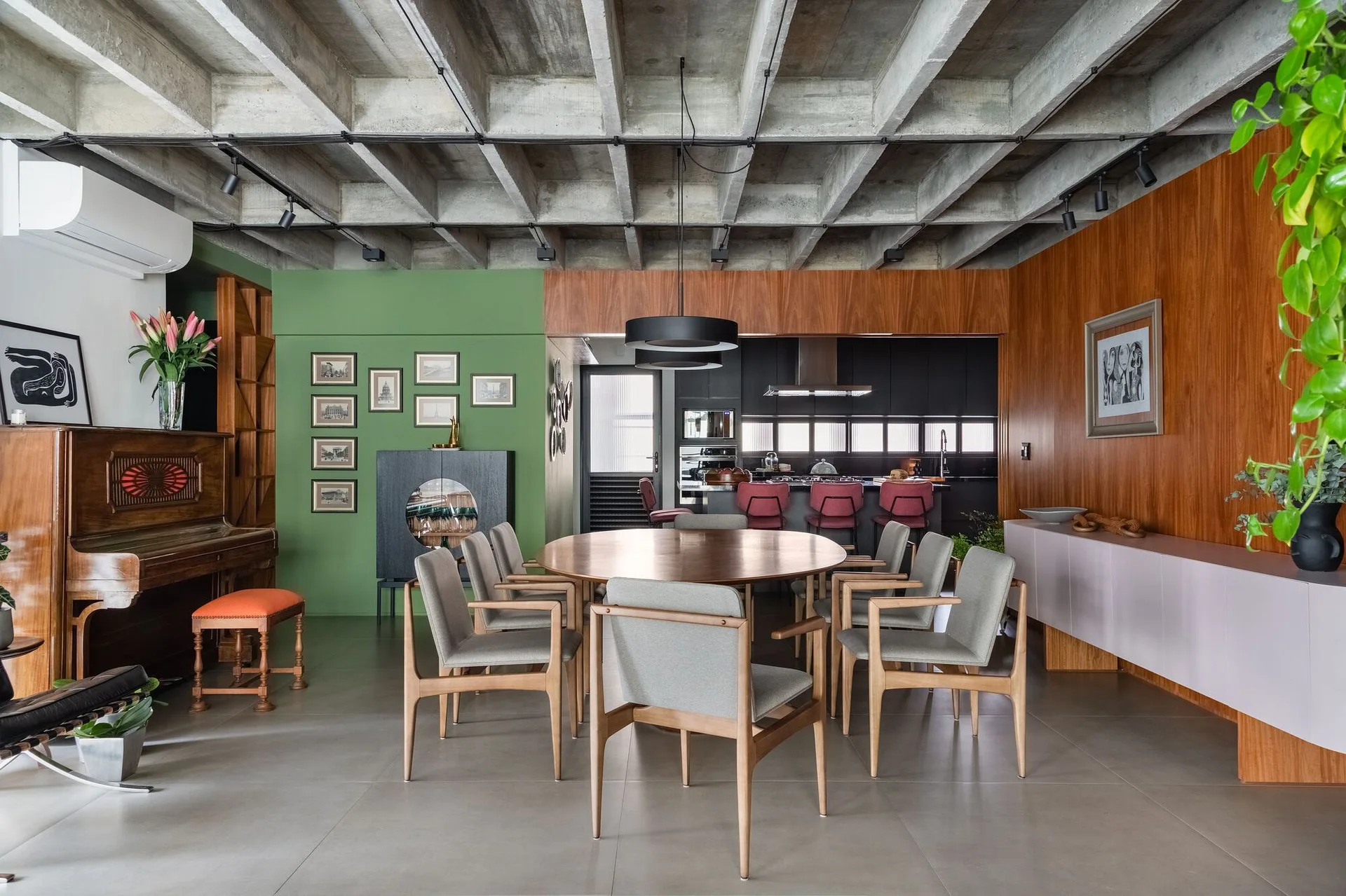 A modern open dining room features a round wooden table, grey upholstered armchairs, an exposed concrete waffle ceiling, and a green accent wall.