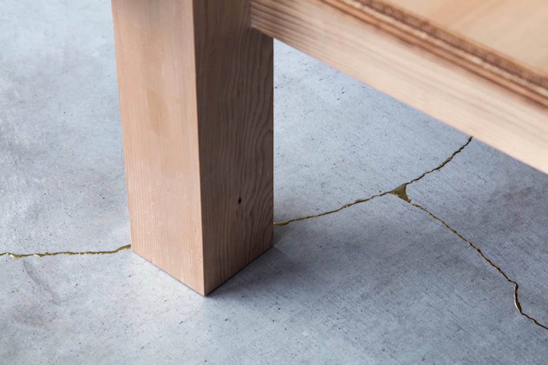 A close-up of a light wood table leg resting on a textured grey concrete floor with cracks repaired with metallic gold fill.