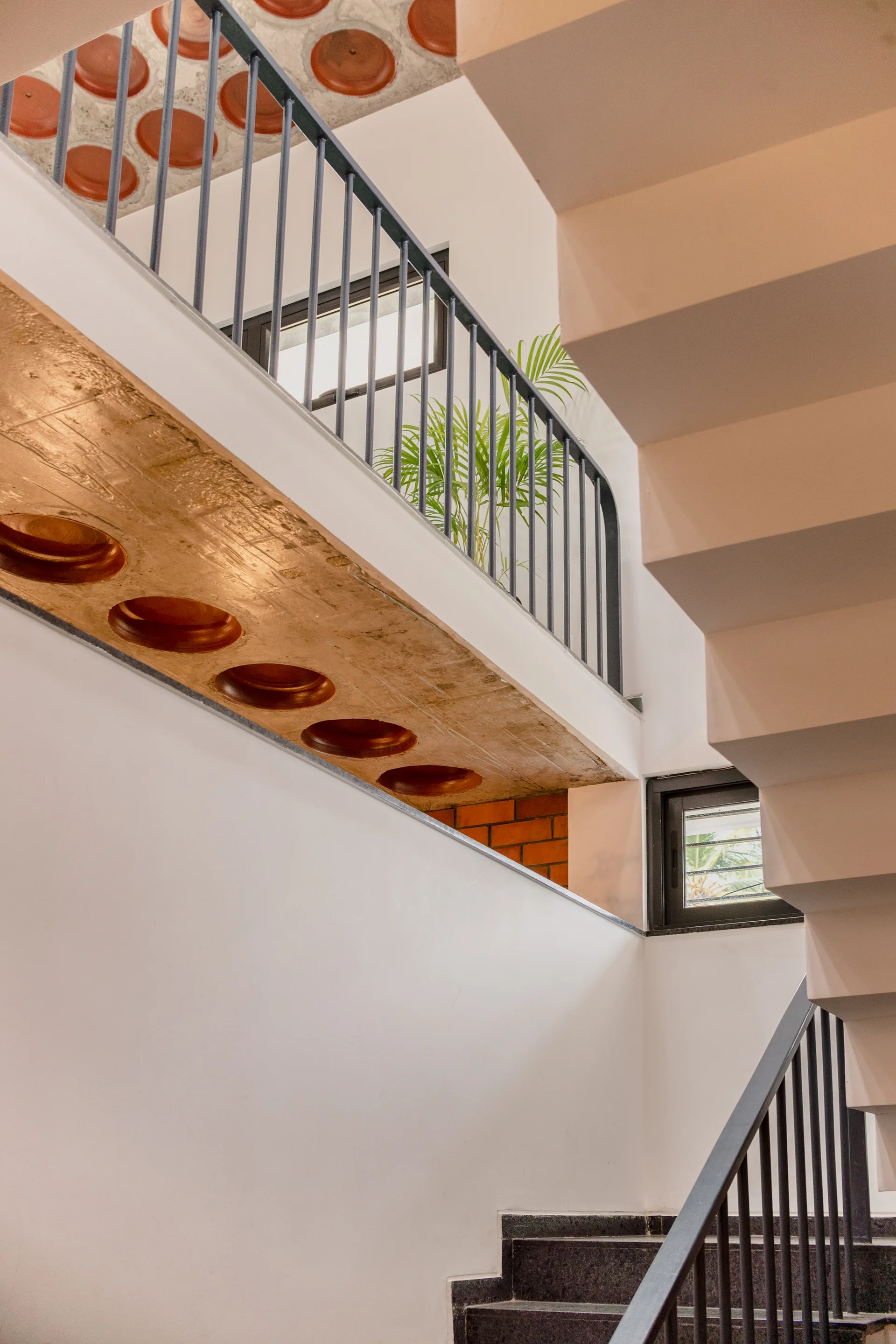 An interior stairwell features white walls, black metal railings, and an exposed concrete ceiling with embedded circular terracotta tiles.