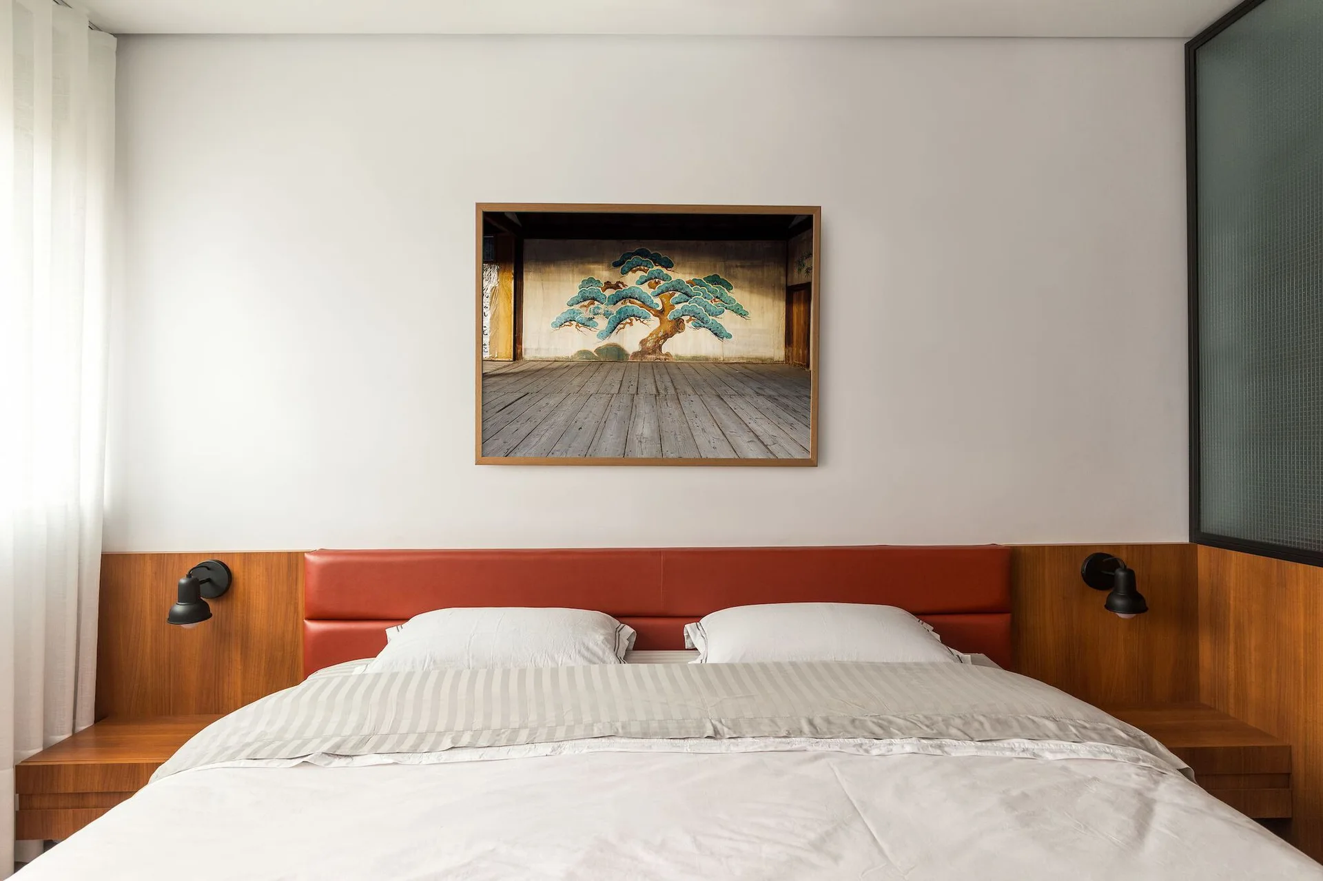 A modern bedroom with a red leather headboard set against horizontal wood paneling, featuring integrated side tables and a framed tree art piece.