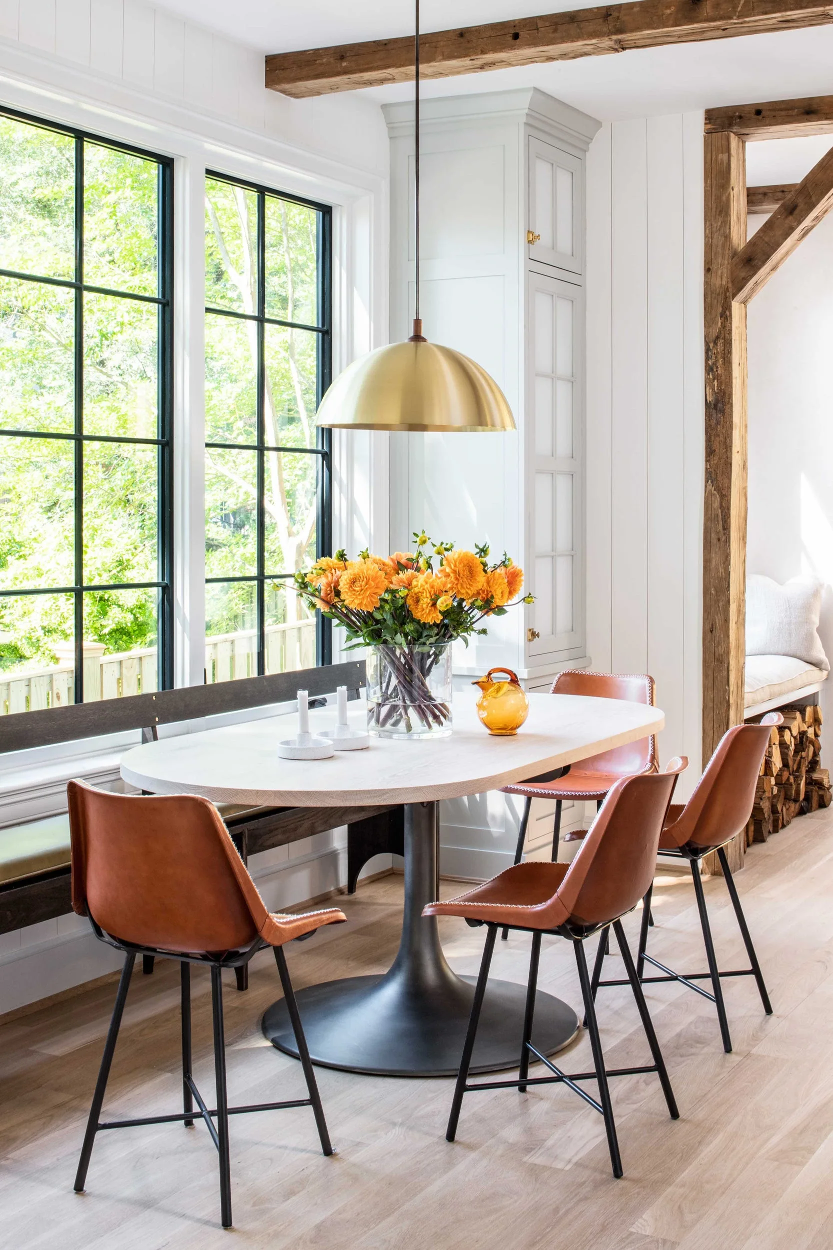 A bright dining area features an oval wood table surrounded by brown leather chairs, a built-in bench under a large grid window, and a brass pendant light.