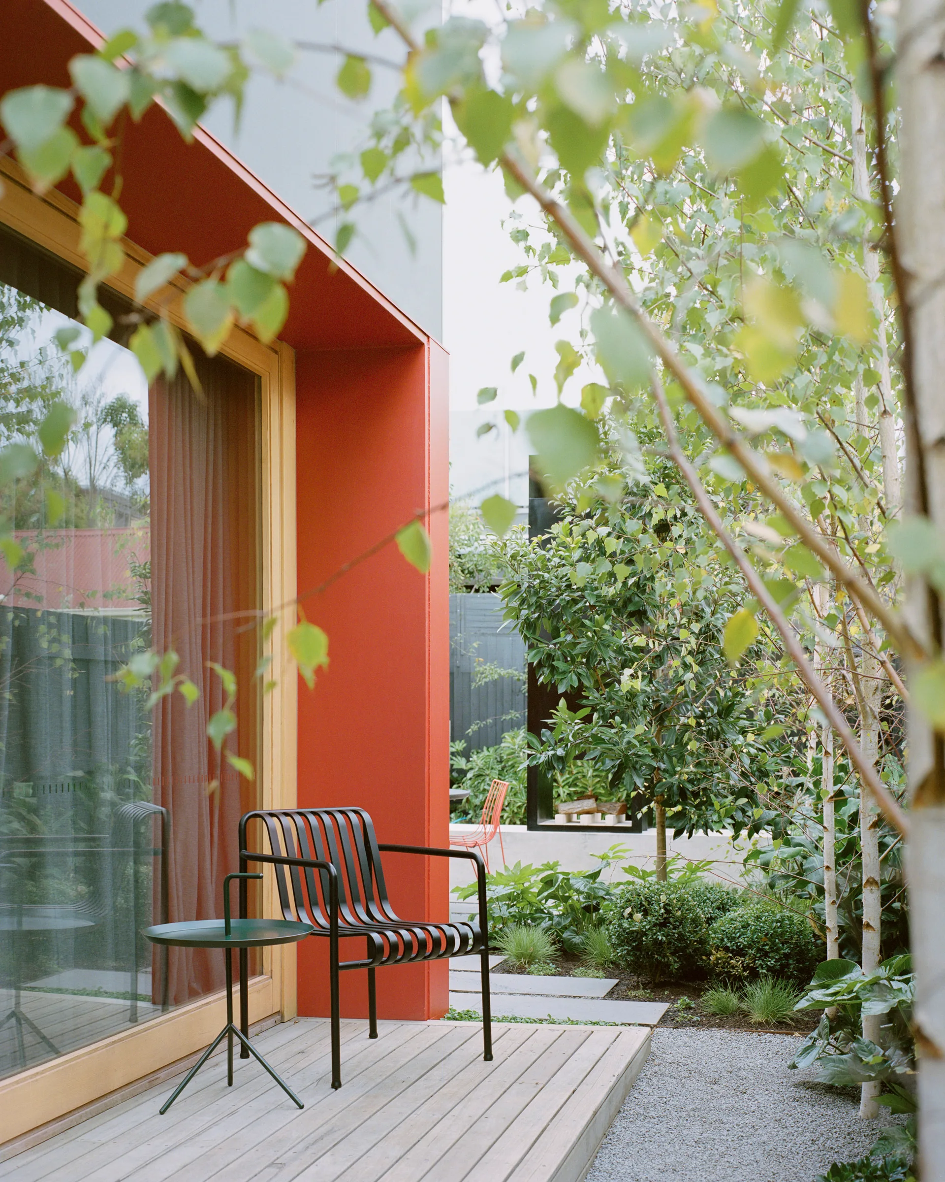 A black metal slat chair and small side table sit on a wooden patio deck next to a bright red facade framing a glass window.