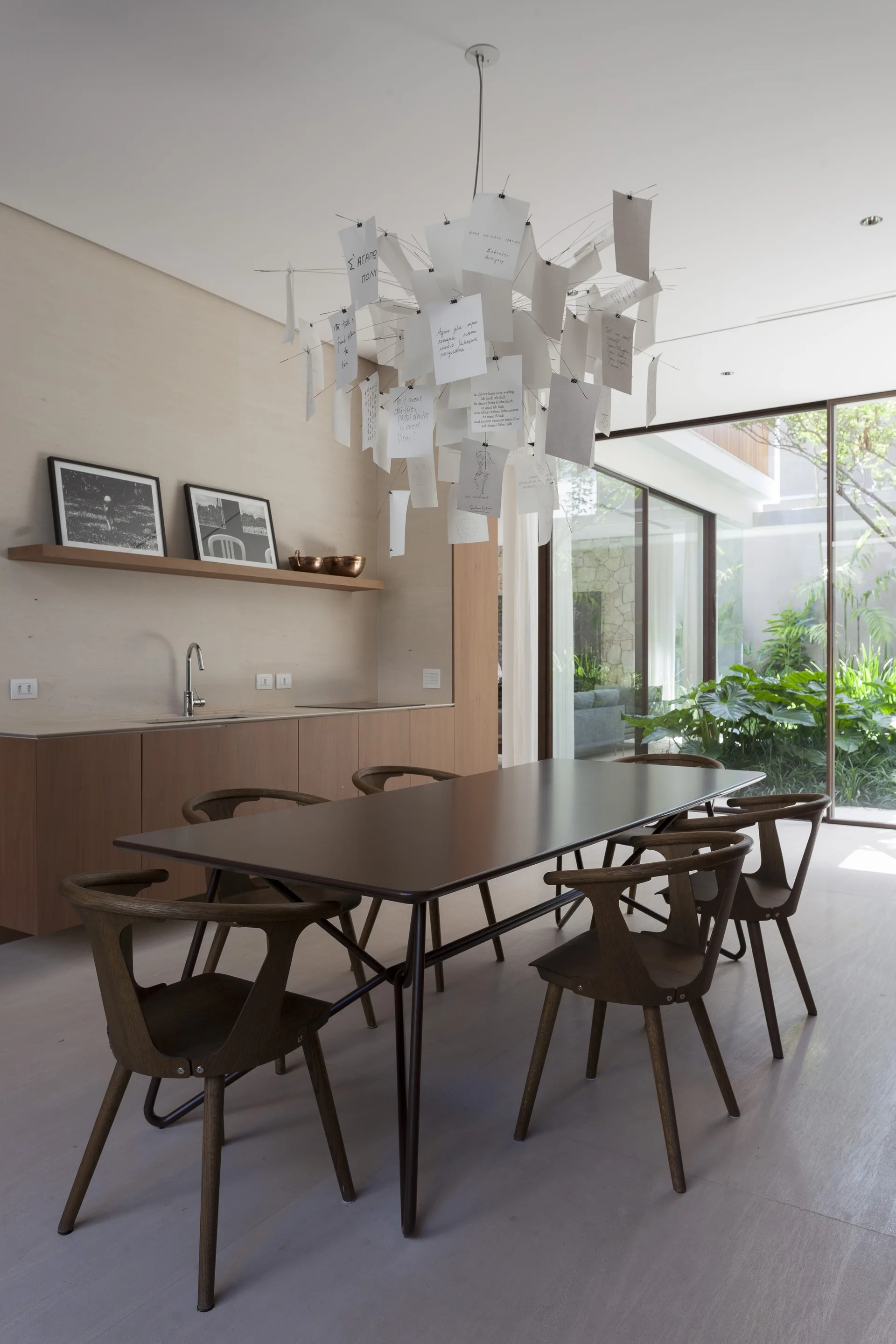 A minimalist dining room features a dark wood table and chairs beneath a paper note chandelier, next to a glass wall showing a lush courtyard.