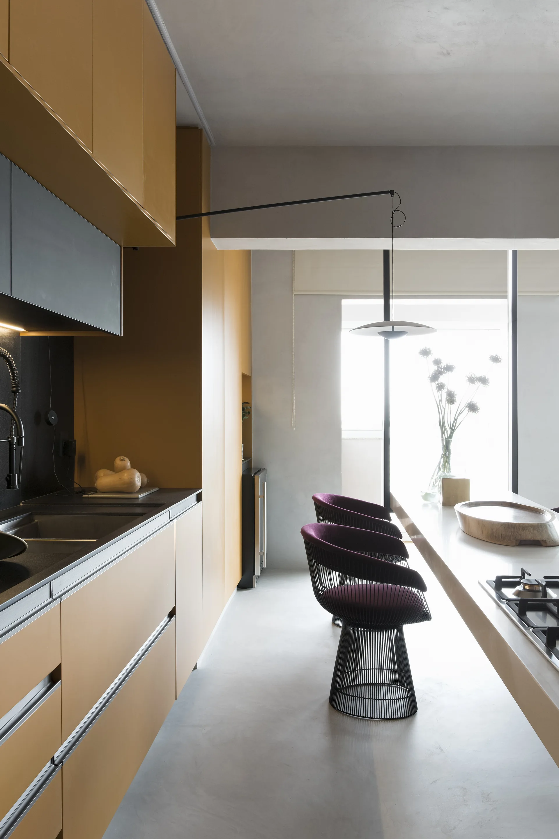 A minimalist kitchen features matte ochre yellow cabinets, a long white island with wire-frame bar stools, and a large window in the background.