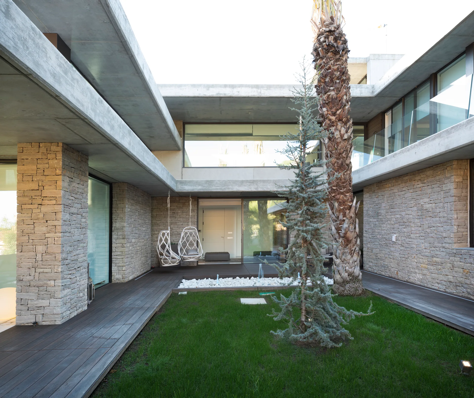 A modern outdoor courtyard features stacked stone walls, wood decking around a small lawn, and hanging chairs beneath a concrete overhang.
