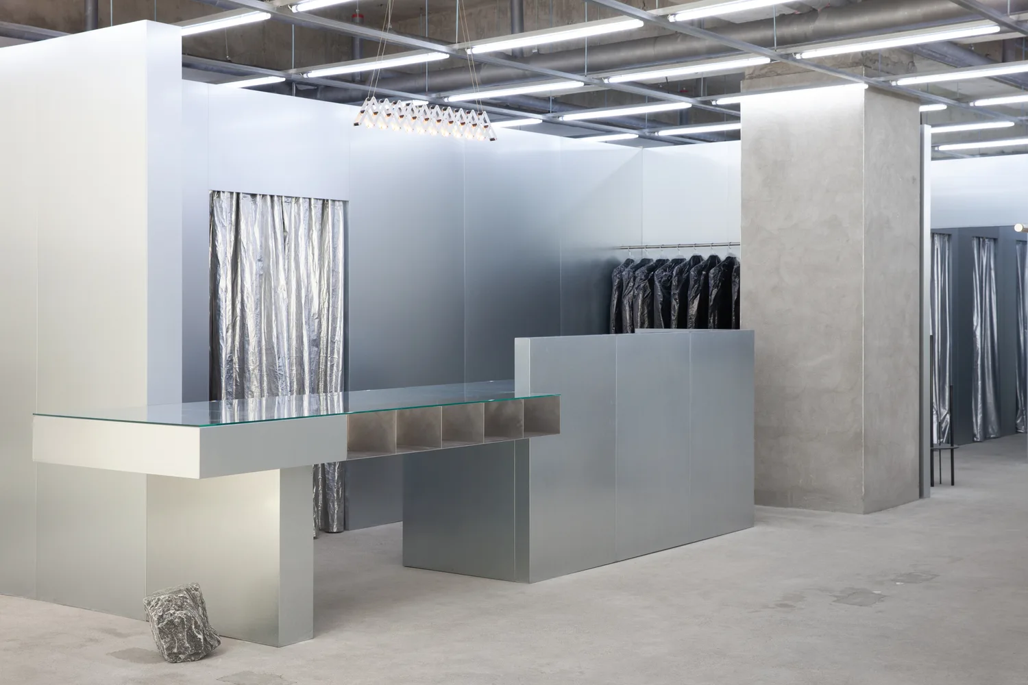 A minimalist commercial interior features silver metallic panel walls, a concrete pillar, and a long glass-top metal display table under a grid ceiling.