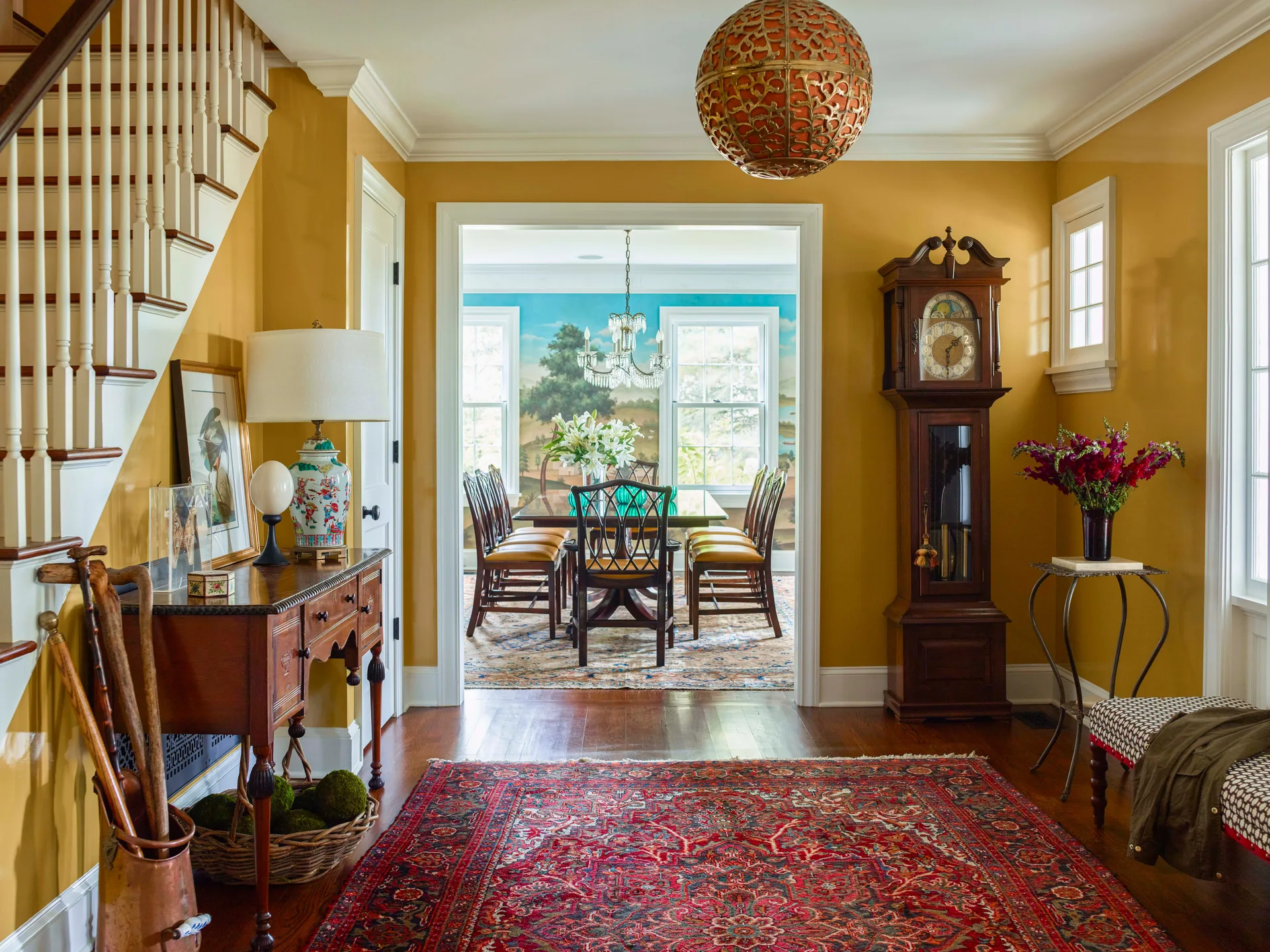 A traditional entryway featuring mustard yellow walls, a wooden console table, a grandfather clock, and a doorway showing a dining room with a scenic mural.