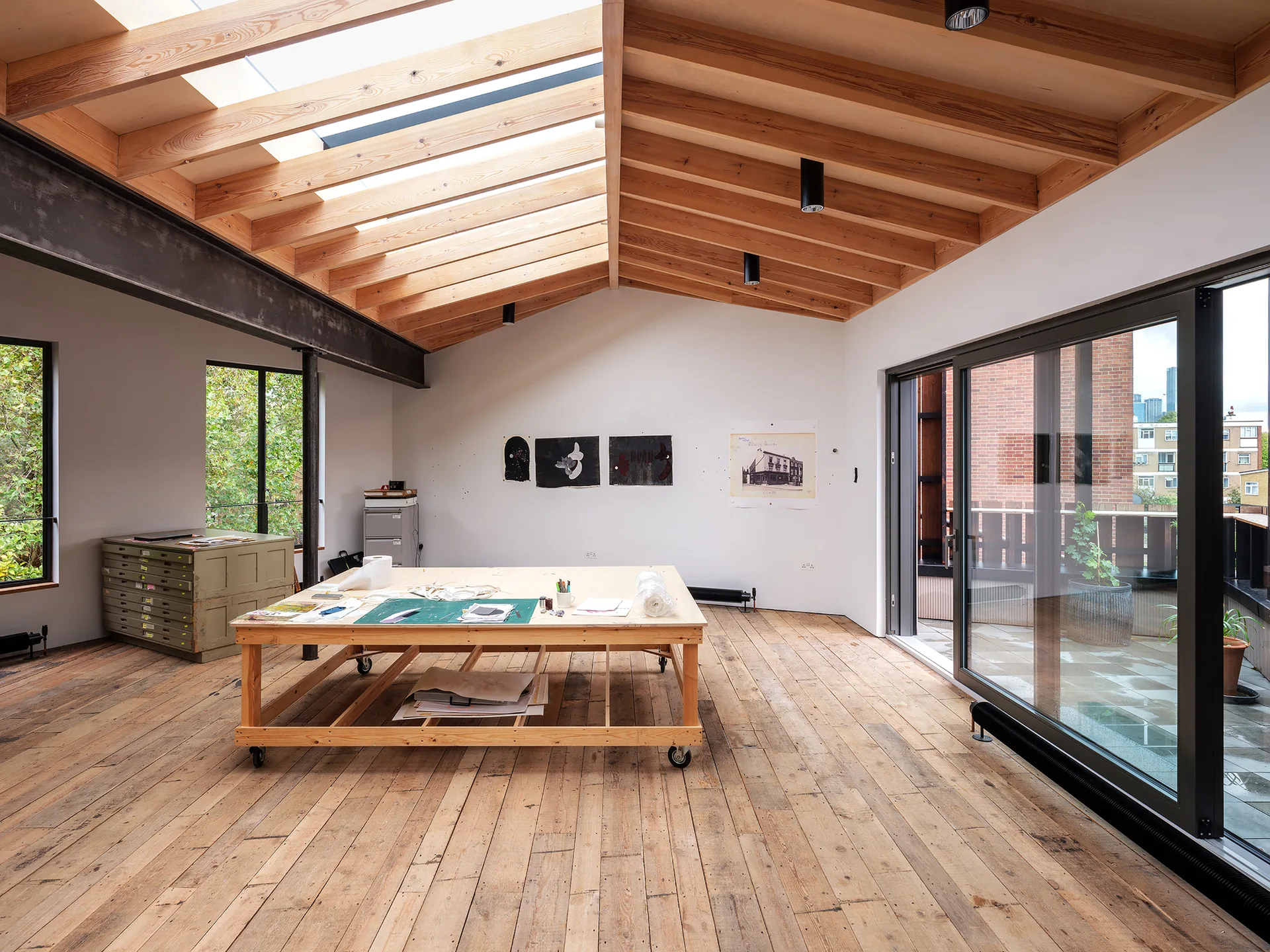 A spacious artist workspace features a central wooden work table on wheels under a vaulted ceiling with exposed timber beams and skylights.