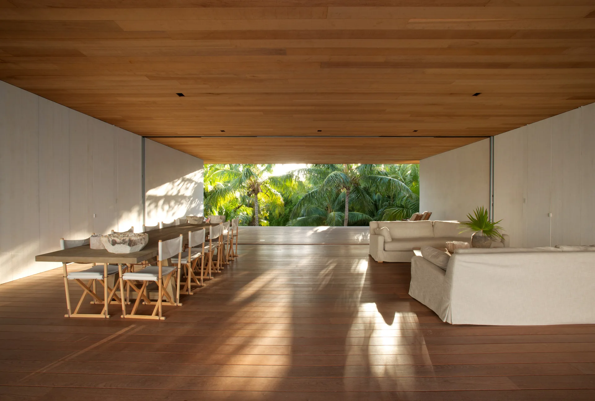An open-air living space featuring a long wooden dining table, canvas chairs, and beige sofas under a matching wood plank ceiling and floor.