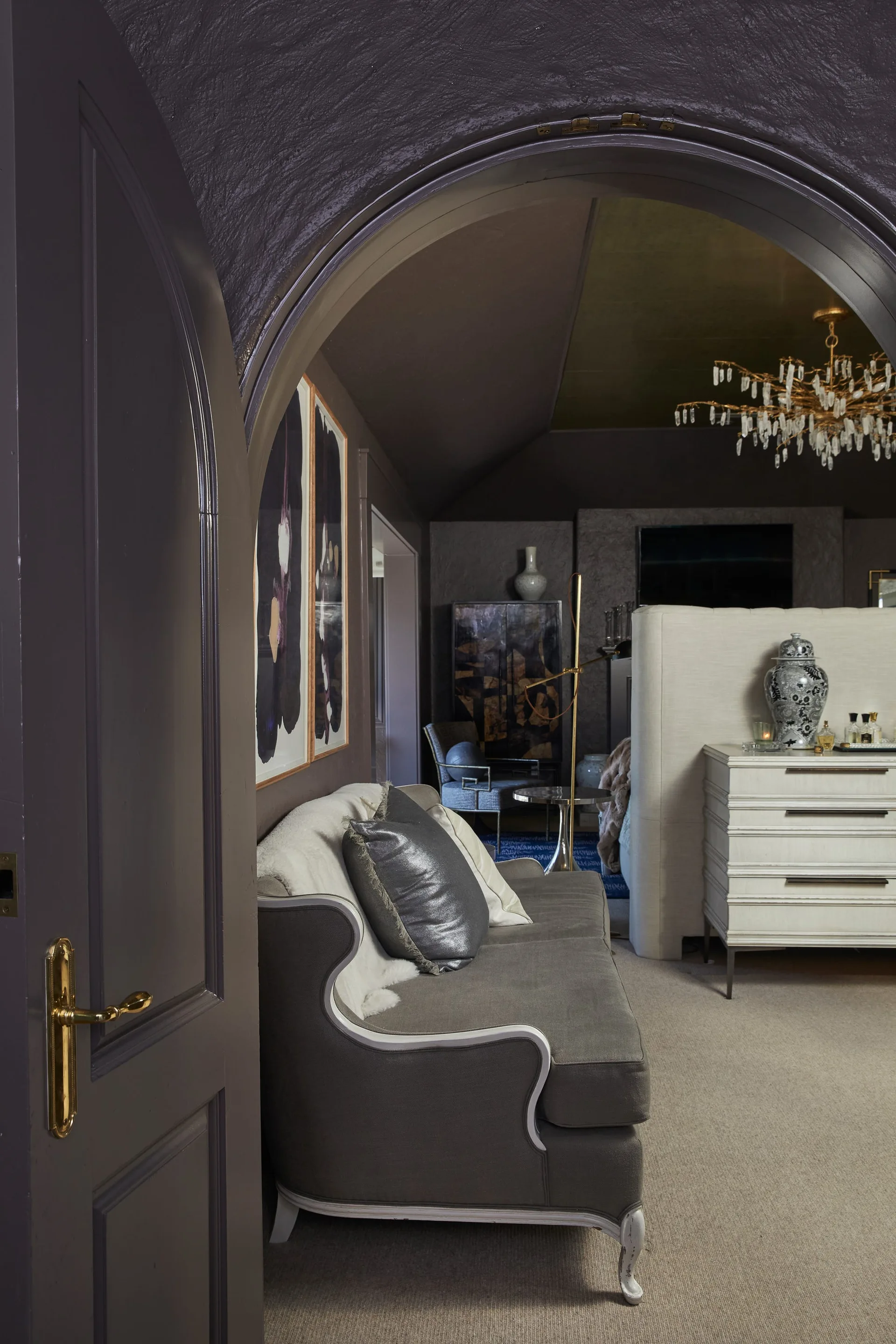 A dark purple arched doorway frames a view into a bedroom featuring a gray sofa, a white dresser, and a gold crystal chandelier.