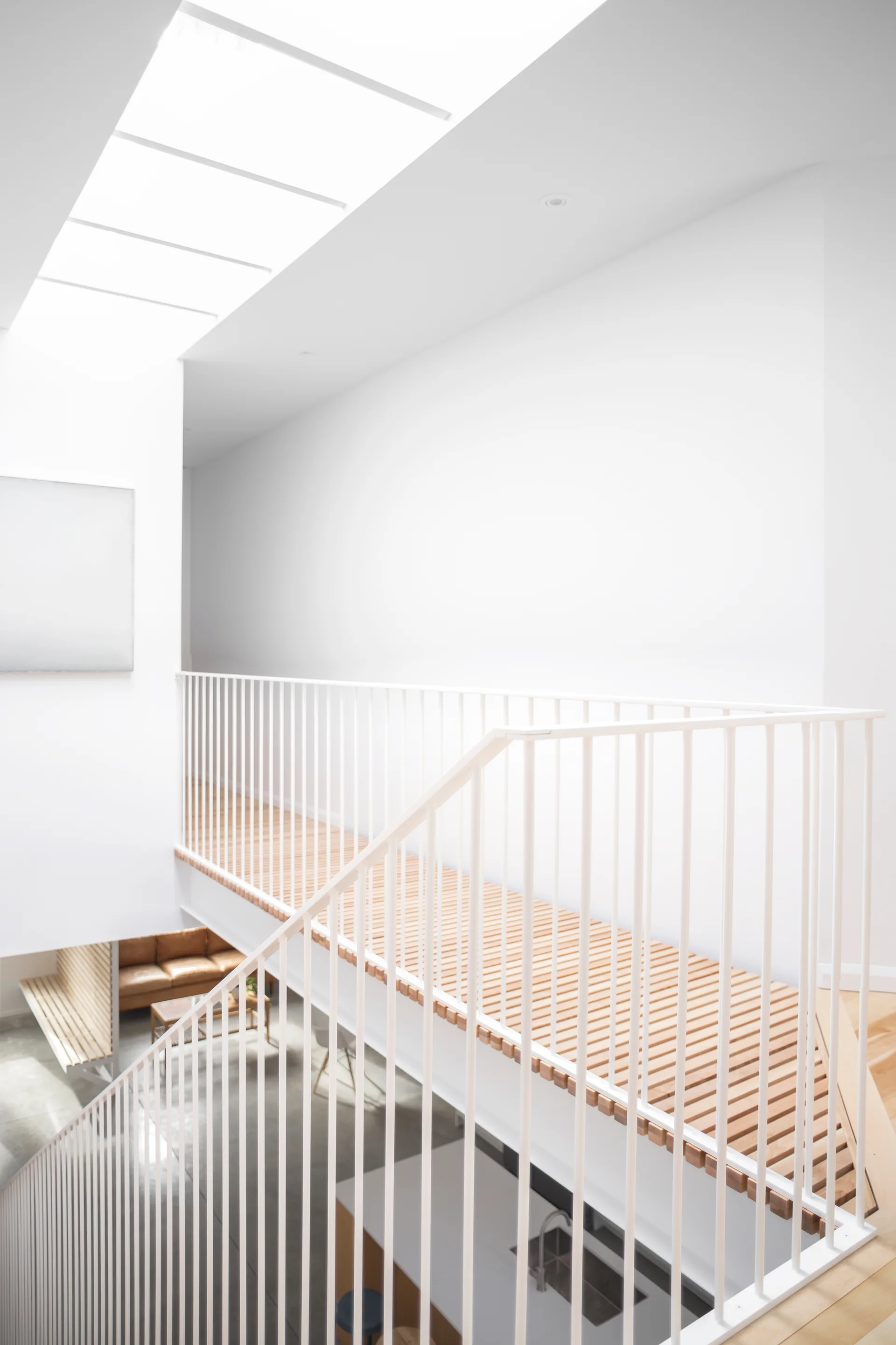 An elevated white metal staircase bridge with slatted wood flooring and vertical railings overlooks a living space lit by a skylight.