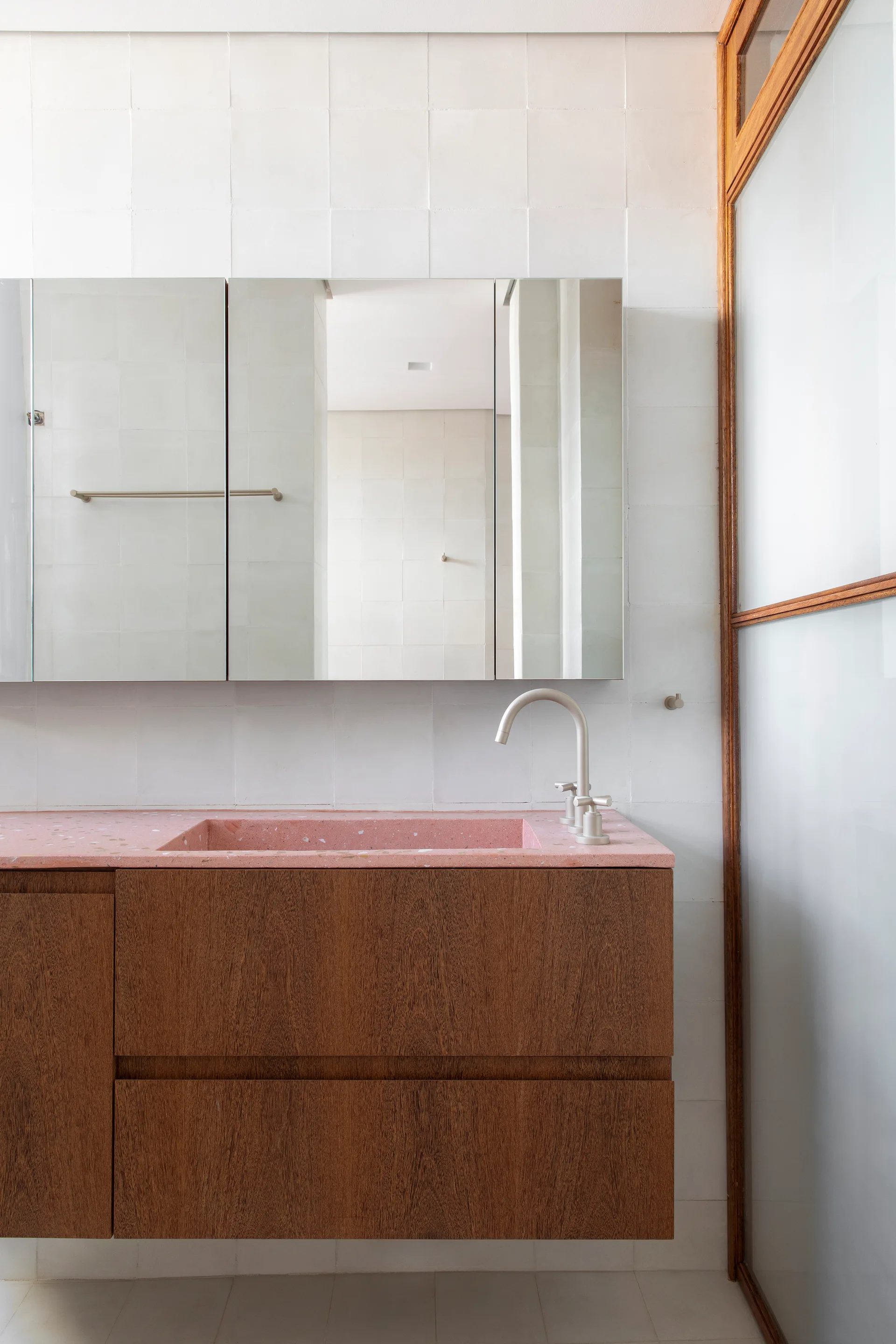 A modern bathroom features a wooden floating vanity with an integrated pink terrazzo countertop and sink against a grid of white square wall tiles.