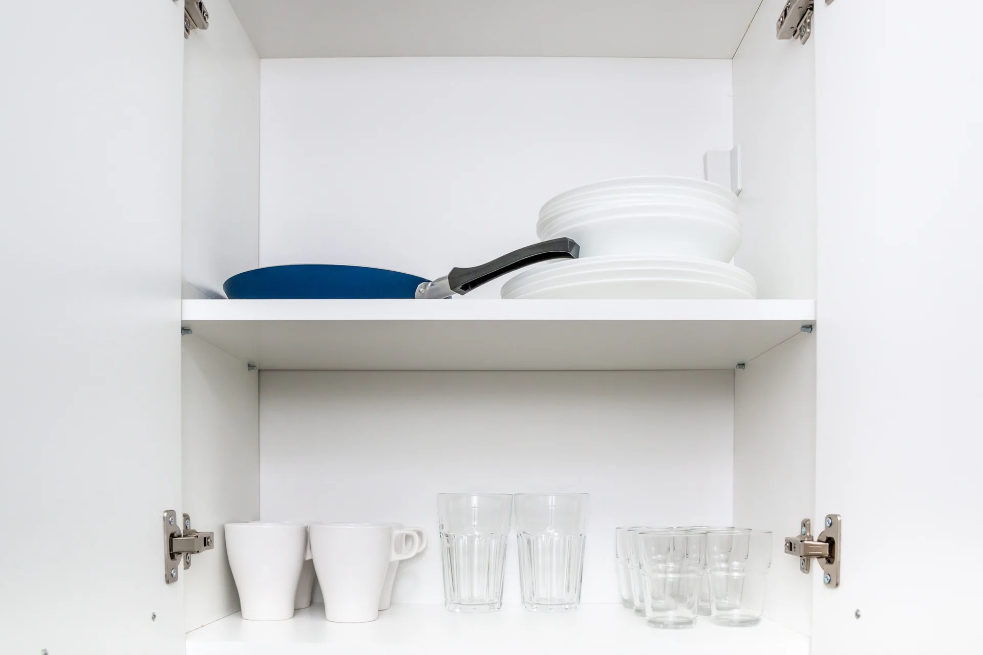 An open white kitchen cabinet featuring neatly organized white mugs, clear drinking glasses, stacked plates, and a blue frying pan.