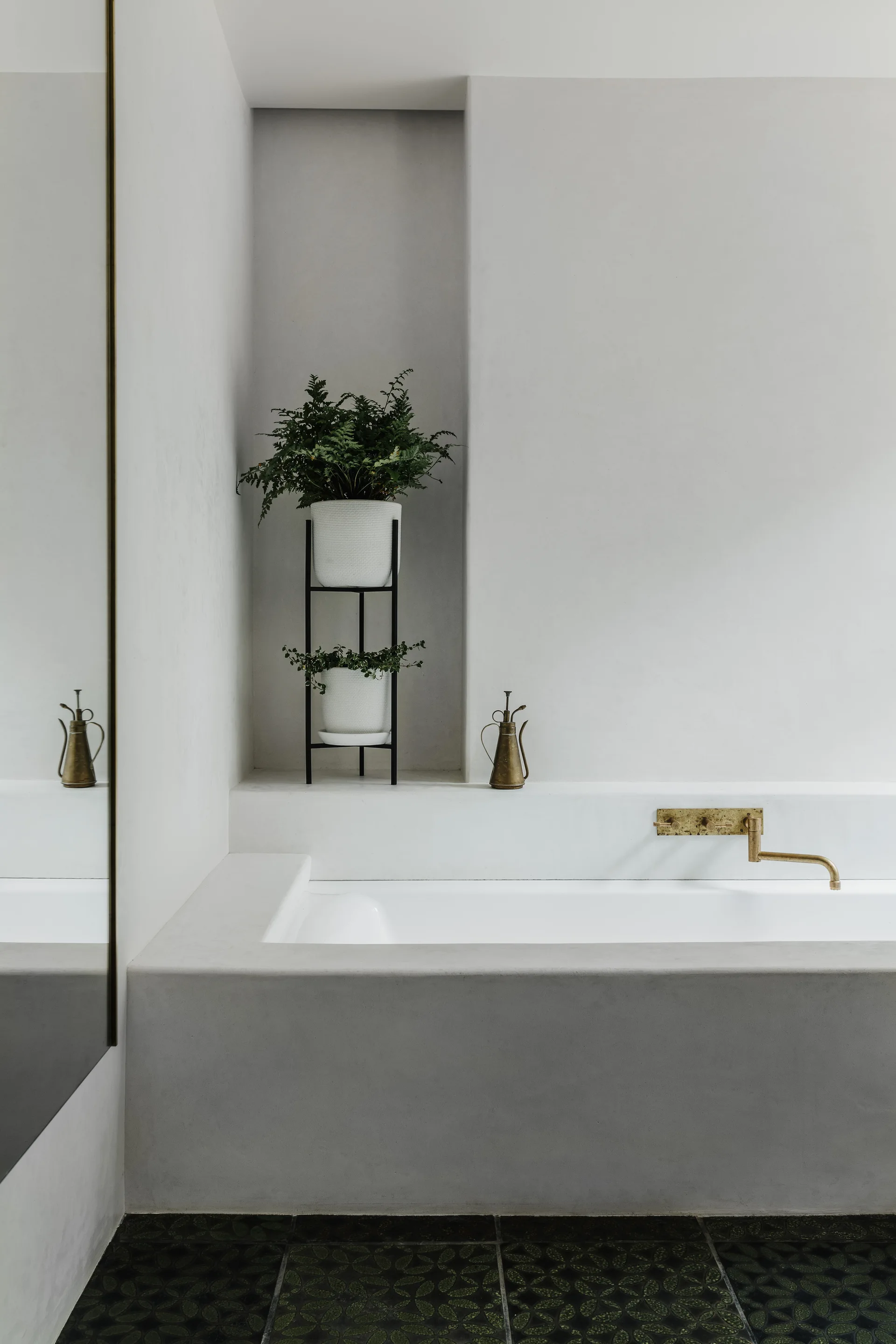 A built-in concrete bathtub features a wall-mounted brass faucet and a recessed wall niche holding green plants in white pots.