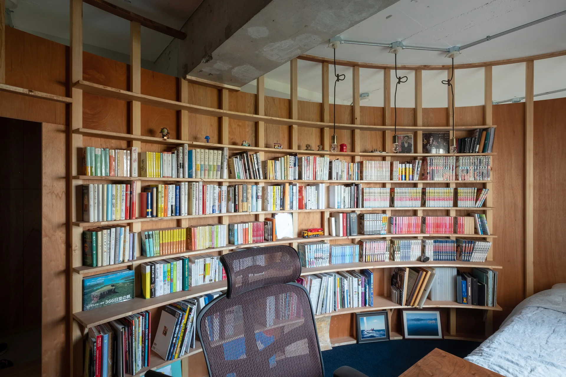 Curved wood shelving filled with books wraps around a curved wall beneath an exposed concrete ceiling with an ergonomic office chair in the foreground.