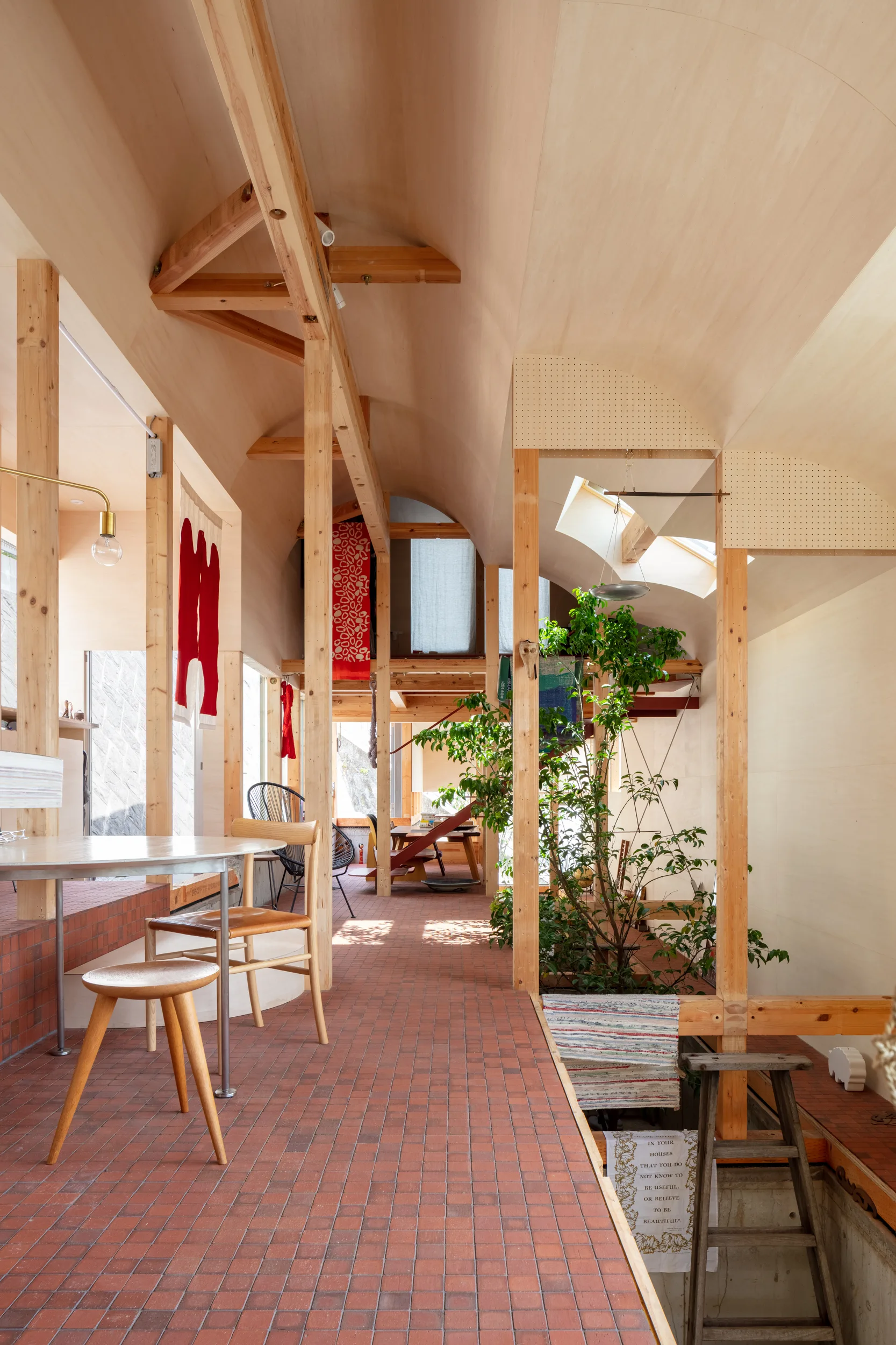 A warm open dining area featuring a vaulted plywood ceiling, exposed wooden timber posts, red brick tile flooring, and a large indoor potted tree.