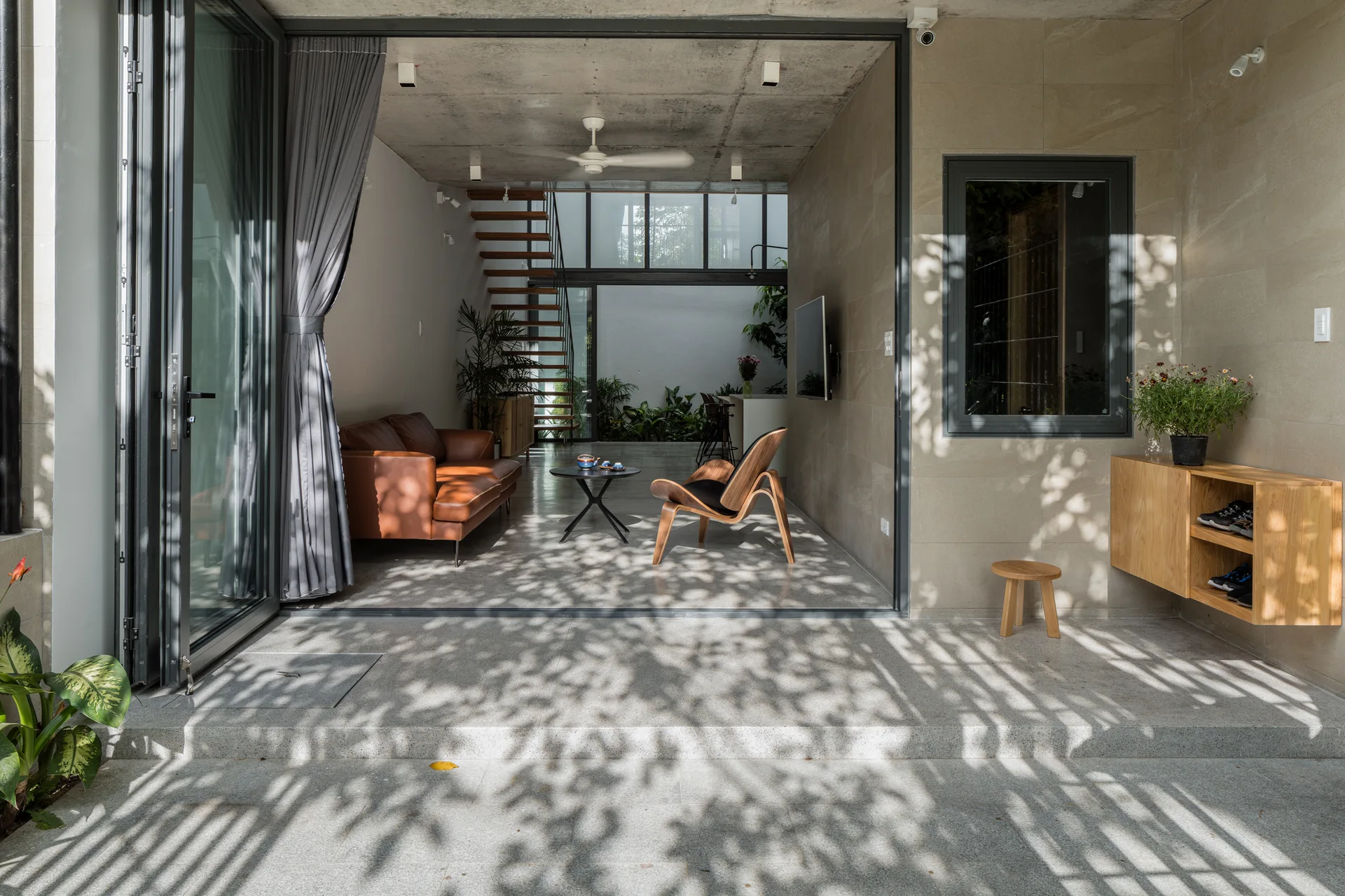 A modern living room with a brown leather sofa and a wooden lounge chair opens to a concrete patio through large black folding glass doors.