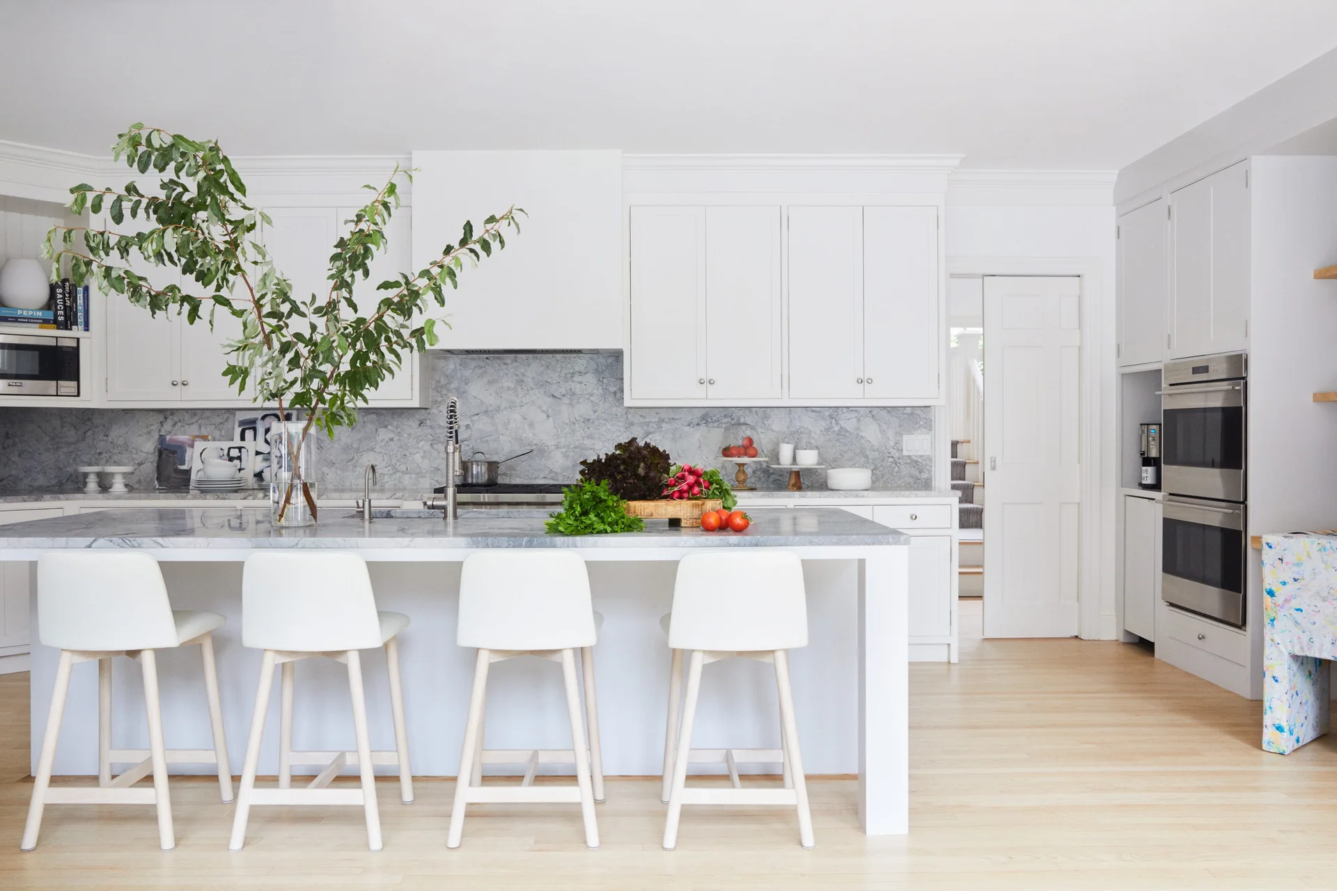 A bright white kitchen features shaker cabinets, a grey veined marble backsplash, and a central island with four white bar stools on light wood floors.