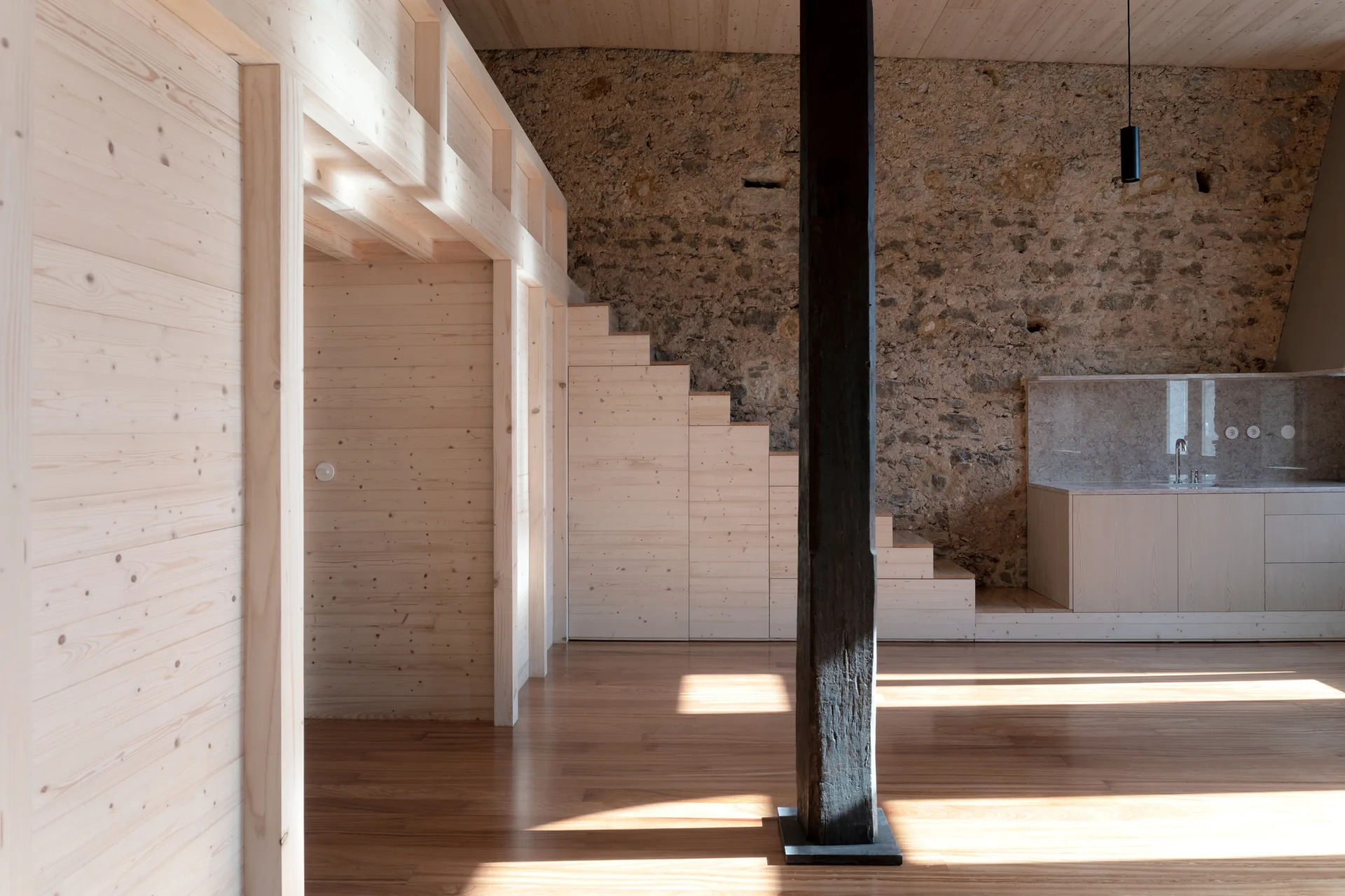 A minimalist interior room features a light wood staircase, integrated storage cabinets, a dark wooden column, and an exposed raw stone wall.