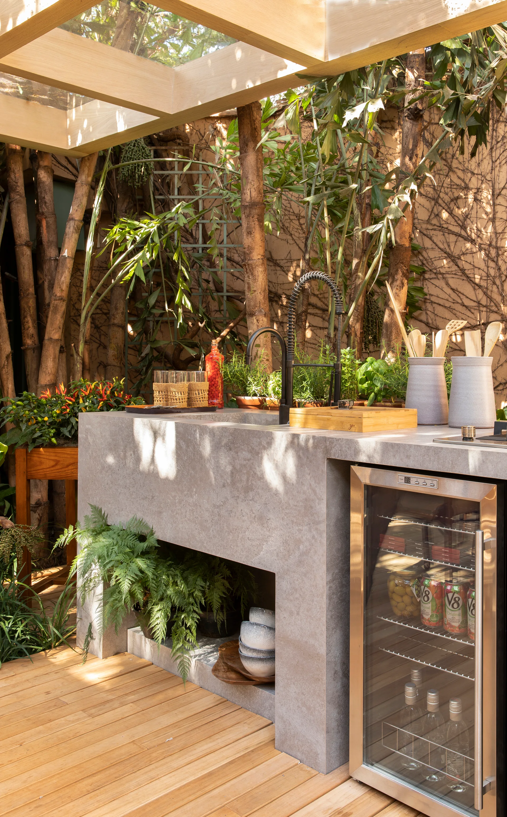A concrete outdoor kitchen island with a built-in glass-front refrigerator and sink sits on a wood deck under a glass-topped pergola.