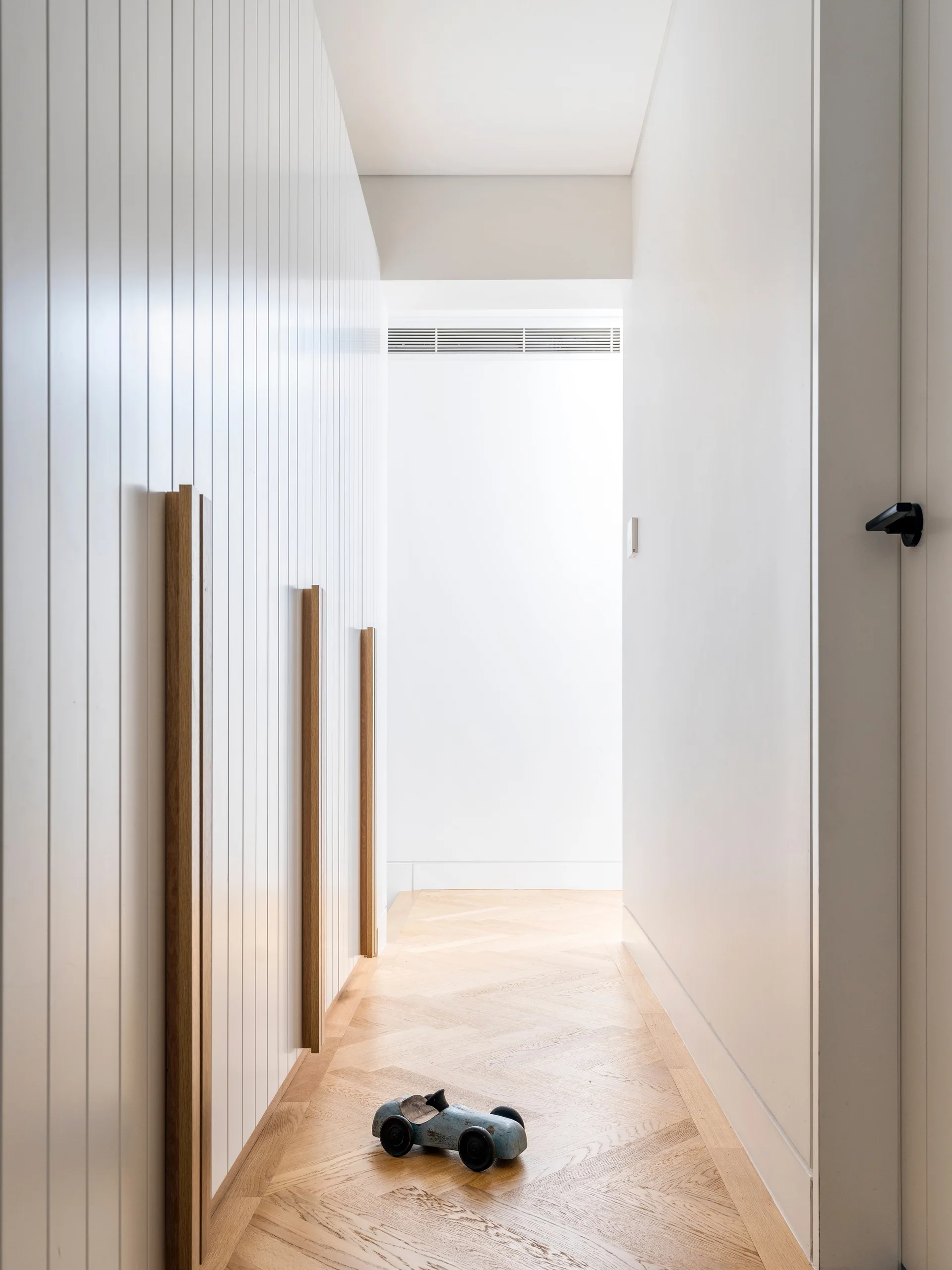 A minimalist white hallway featuring vertical wall paneling, long wood handles, and light-colored herringbone parquet flooring.
