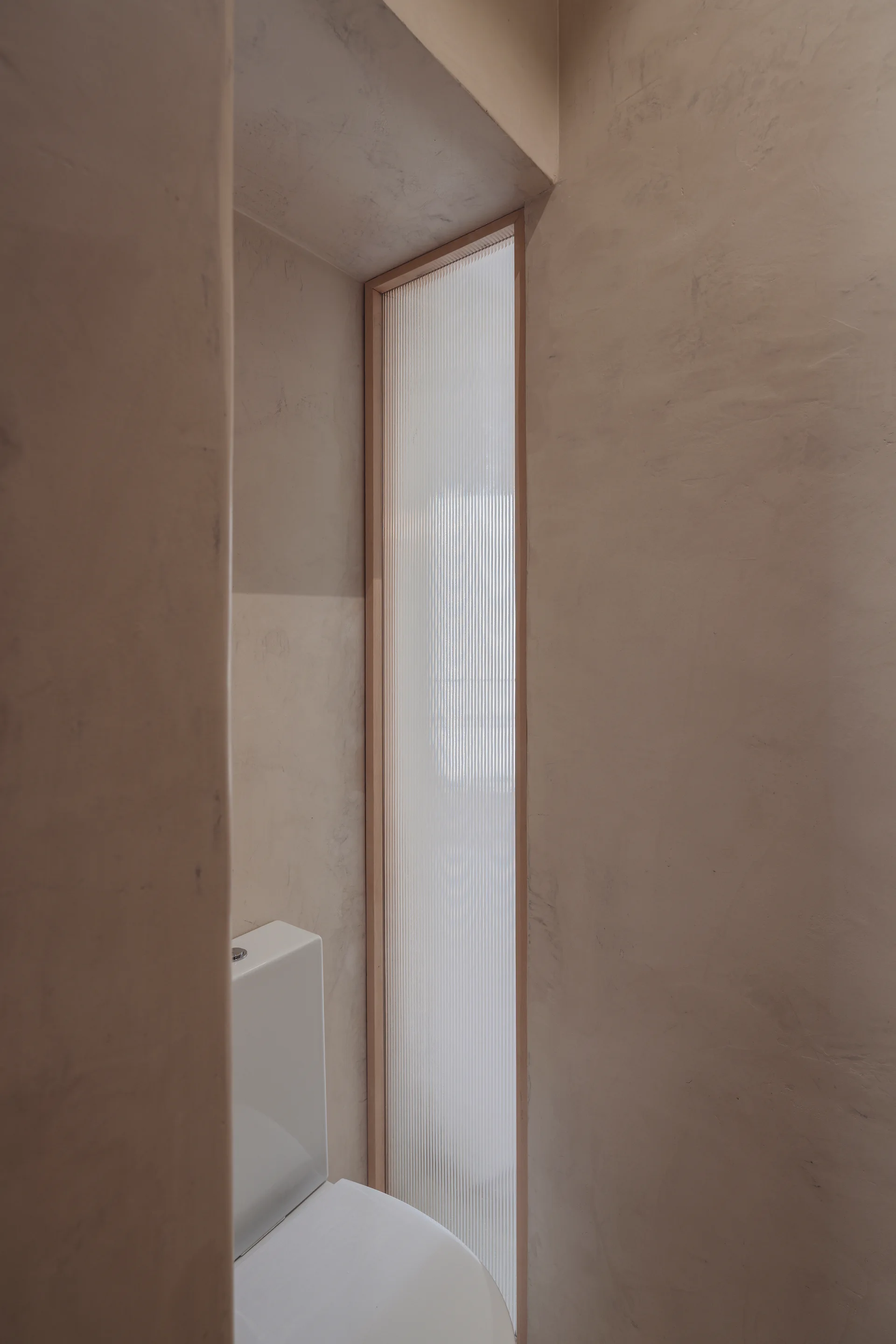 A white ceramic toilet sits in a narrow bathroom niche featuring textured beige plaster walls and a vertical fluted glass window panel.