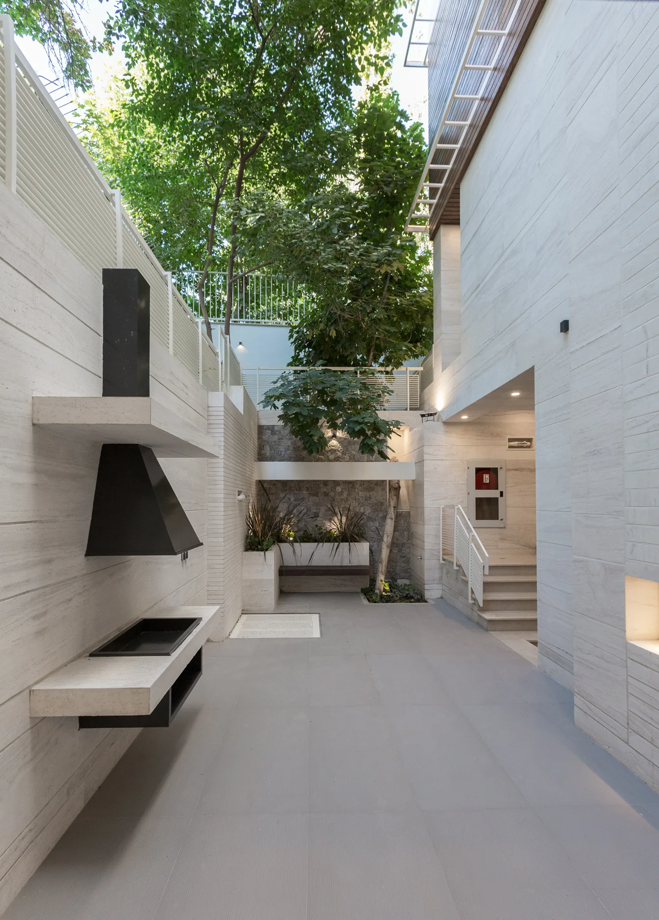 A modern outdoor courtyard featuring beige stone-clad walls, a minimalist black wall-mounted fireplace, a gray tiled floor, and an integrated tree.