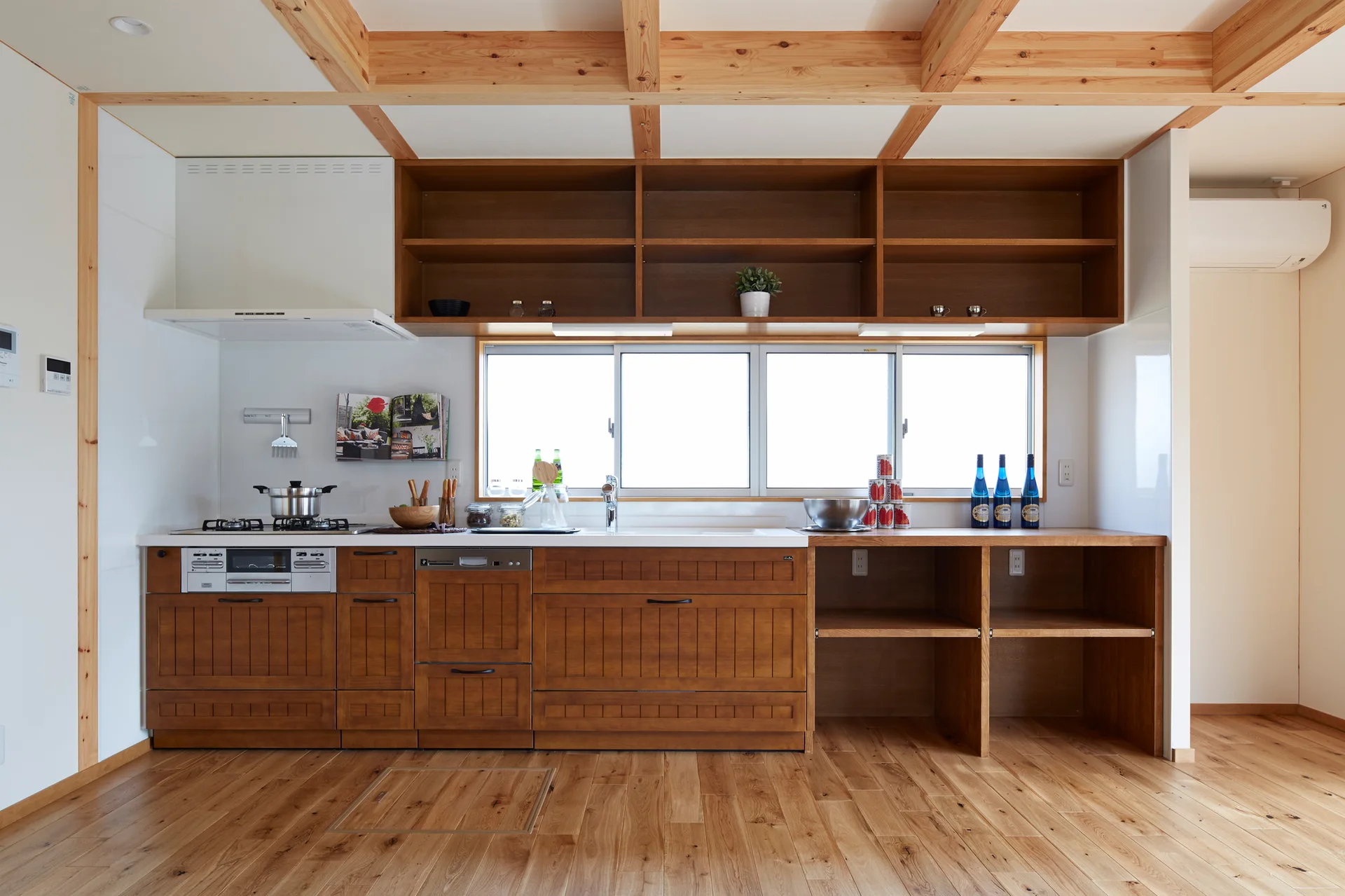 A bright kitchen features warm wood cabinetry, matching open shelving over a central window, and exposed timber ceiling beams.