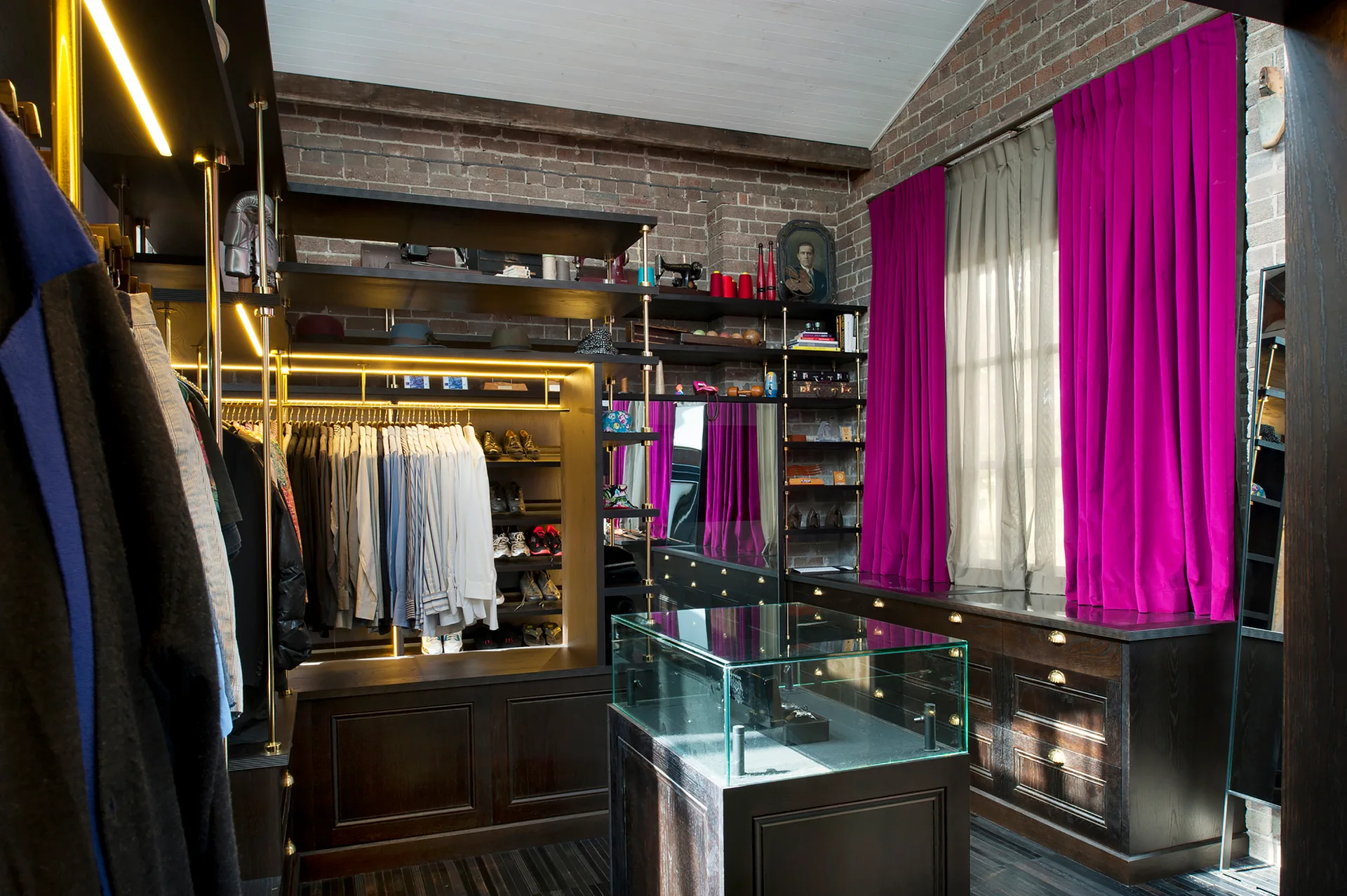 A walk-in closet features dark wood storage cabinetry, exposed brick walls, magenta curtains, and a central glass jewelry display island.