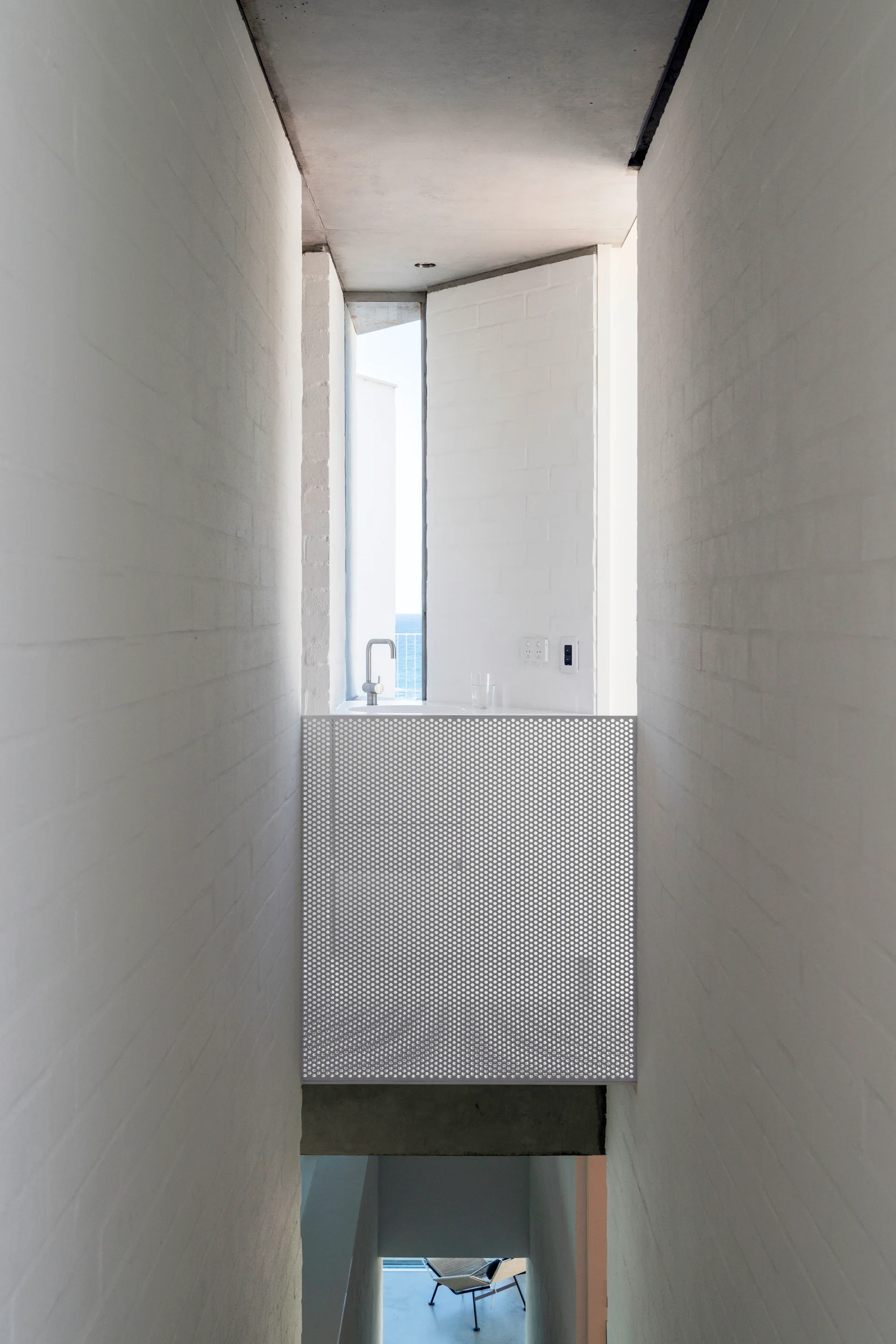 A narrow bathroom hallway with painted white brick walls features a silver faucet behind a grey perforated metal privacy screen.