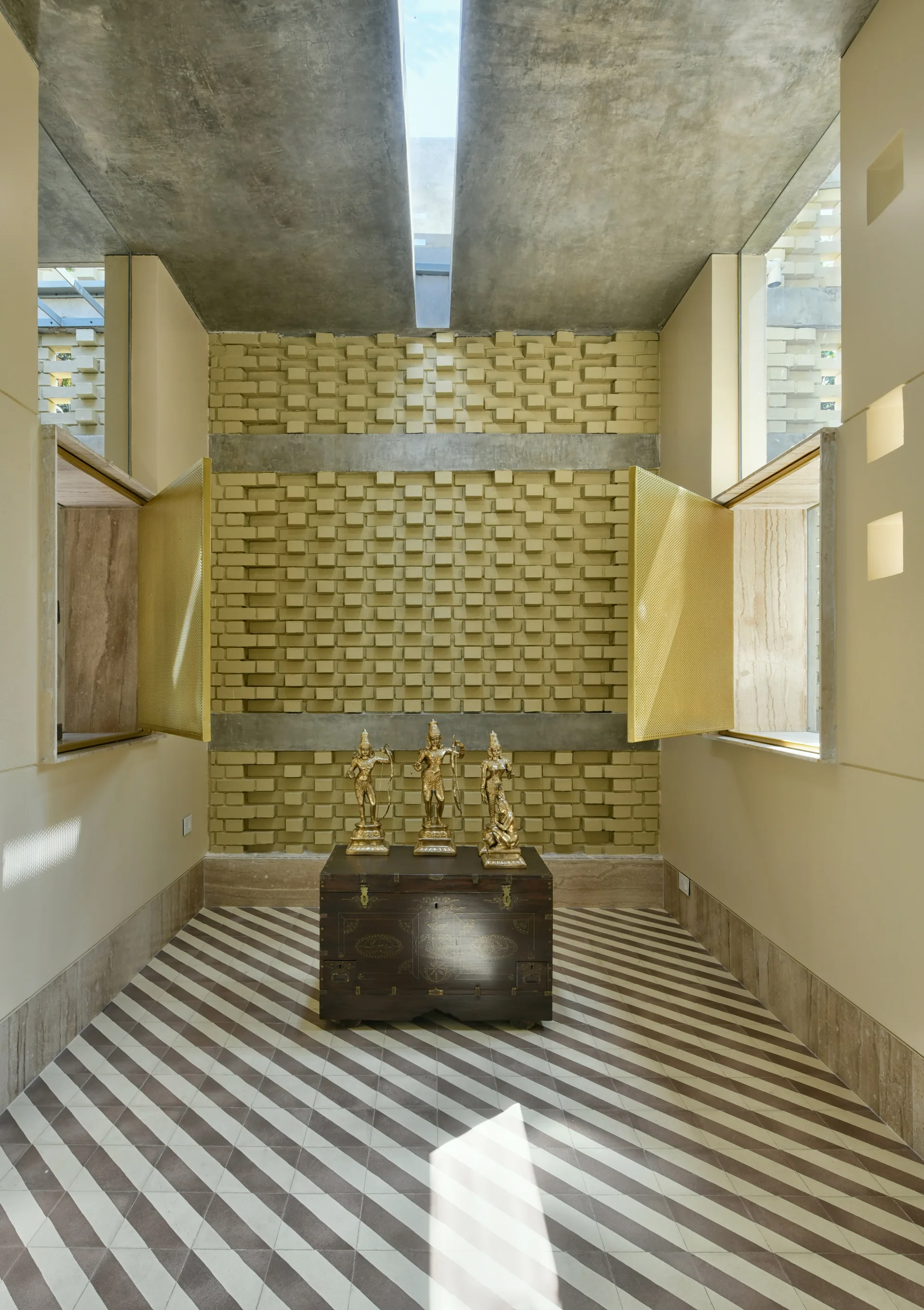 A meditative alcove features three gold statues on a dark wood chest set against a textured yellow brick wall and diagonal striped floor tiles.