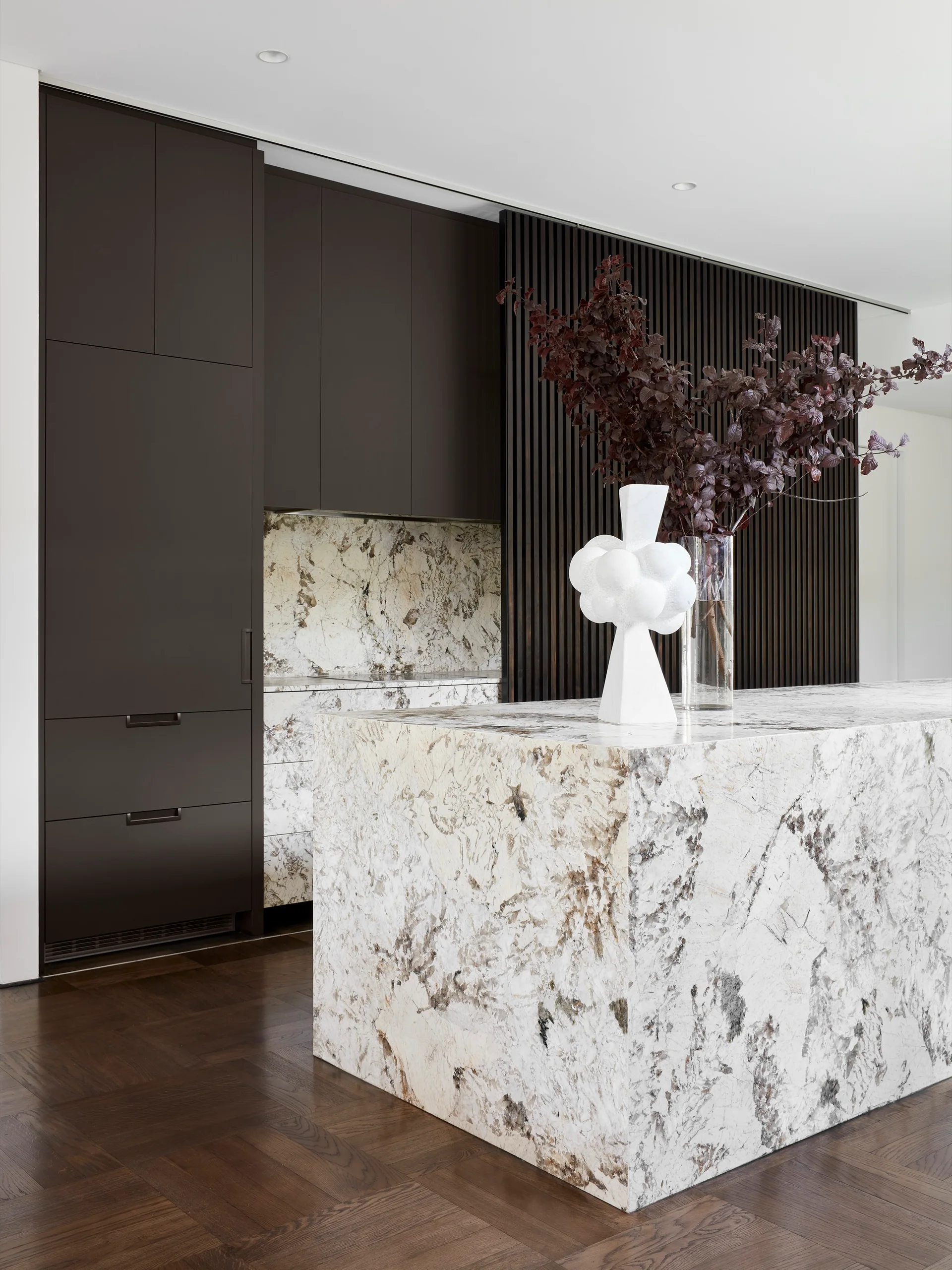 A modern kitchen featuring a heavily veined marble waterfall island, matching backsplash, dark brown flat-panel cabinets, and a vertical wood slat wall.