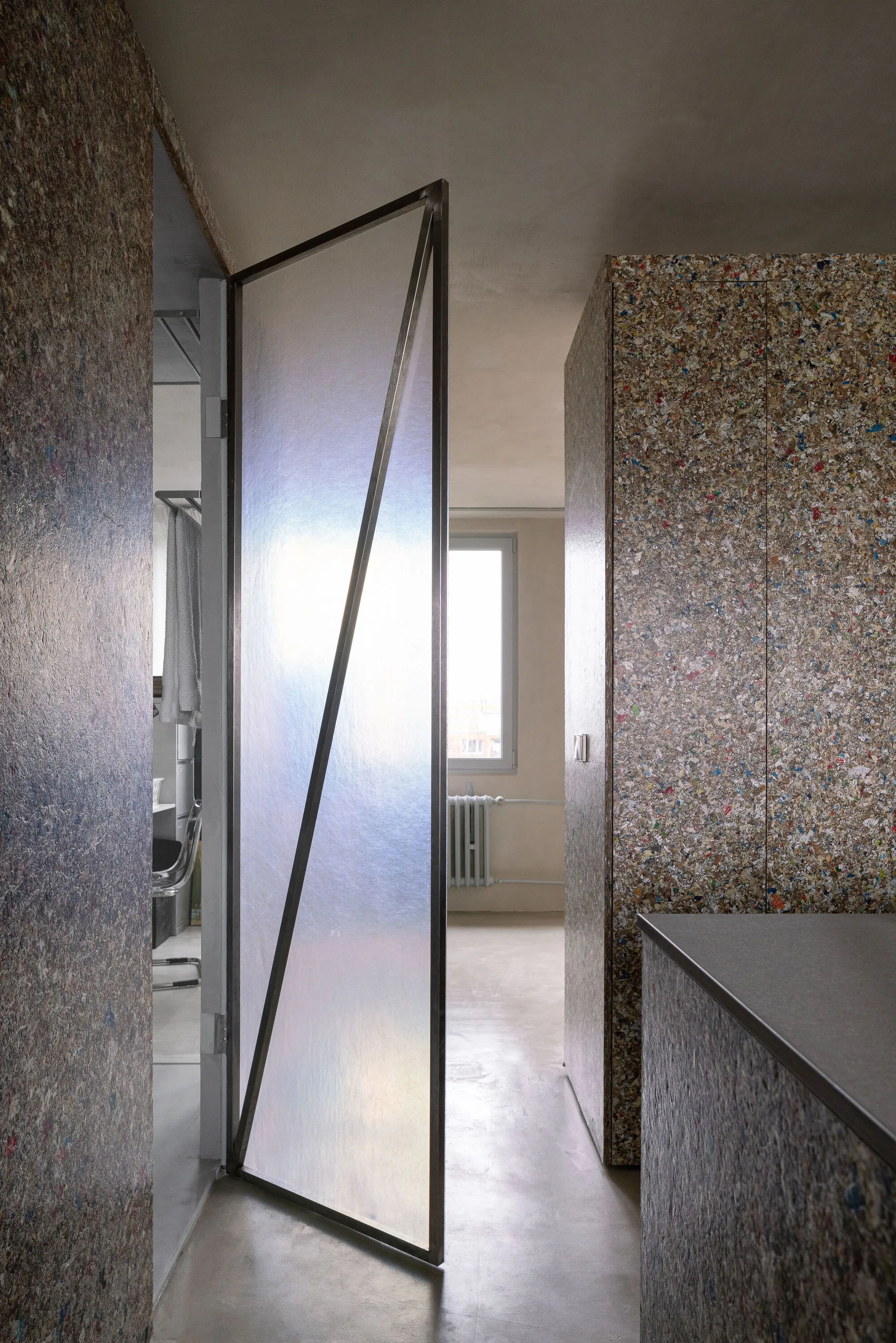 An interior space features a pivoting translucent glass door set against walls and cabinets clad in speckled, multi-colored recycled composite paneling.