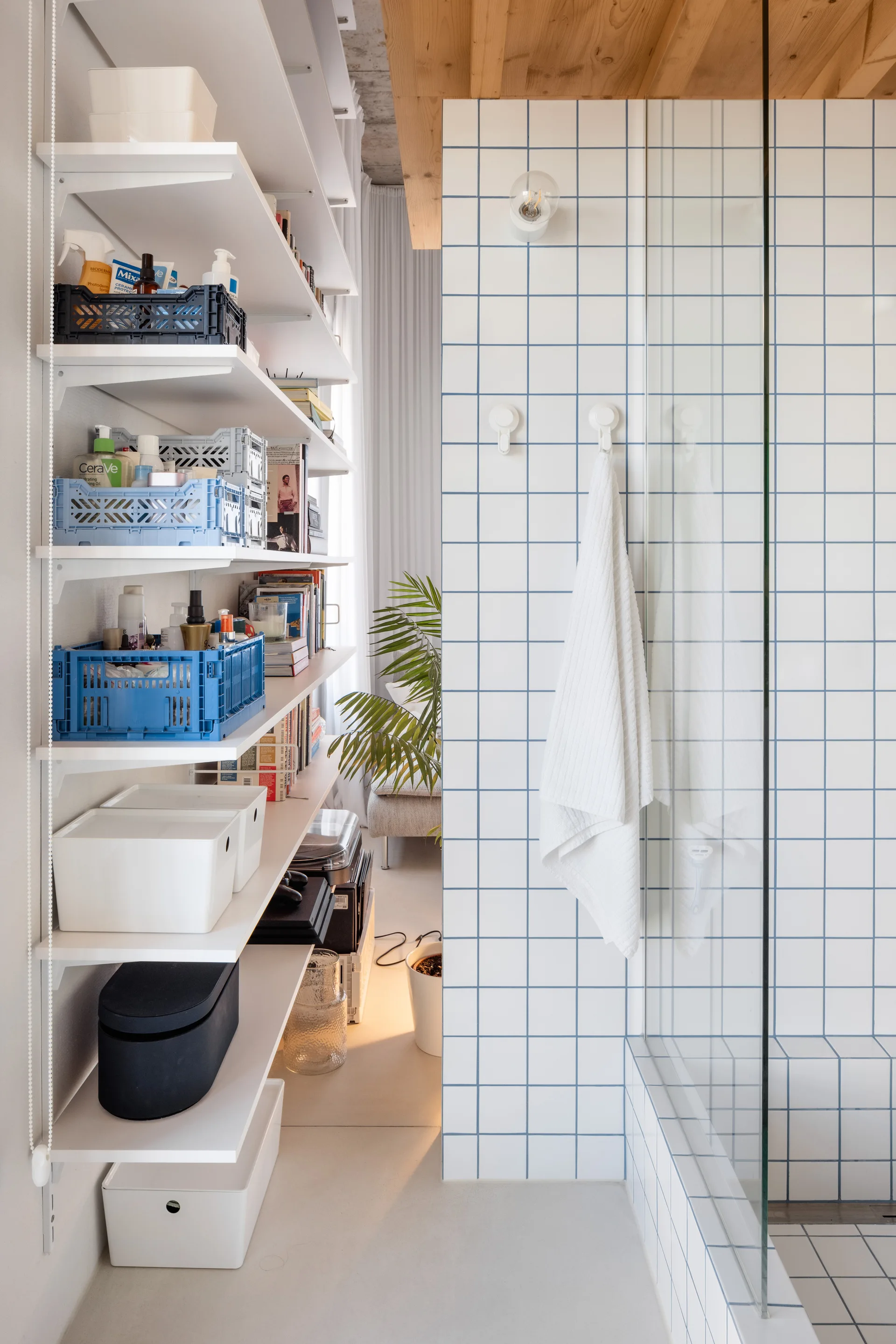 A modern bathroom features a white tiled shower with blue grout lines next to white wall shelves holding blue storage crates and toiletries.