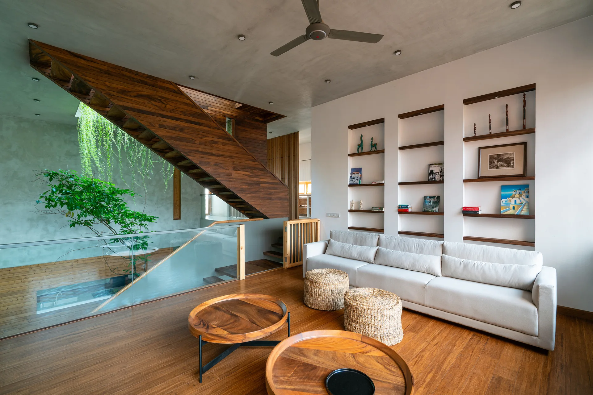 A modern living room featuring a white sofa, round wooden coffee tables, a wooden staircase, and an open atrium with a green tree.
