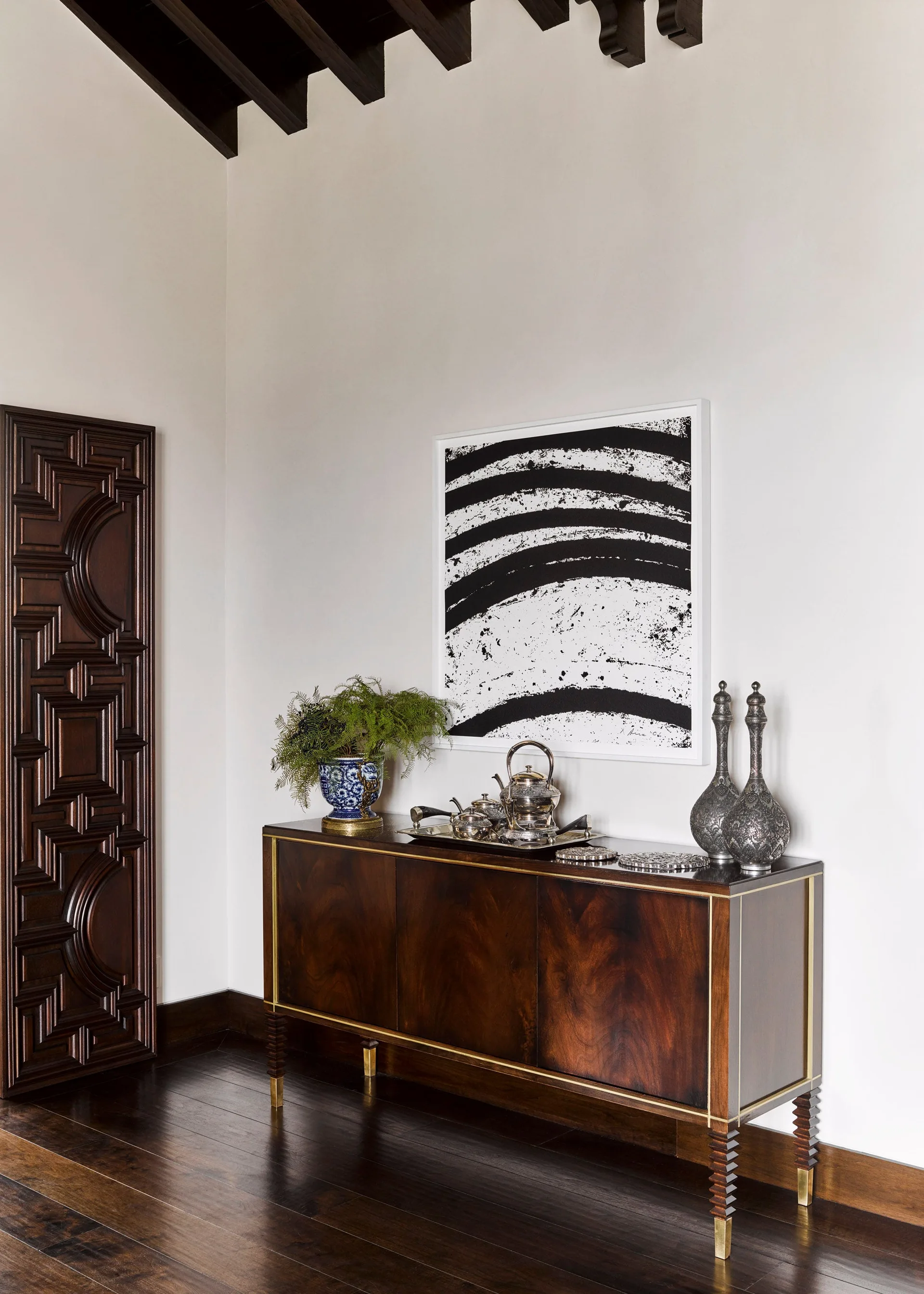 A dark wood console table with brass details stands against a white wall below a black and white abstract painting, next to a carved wooden door.