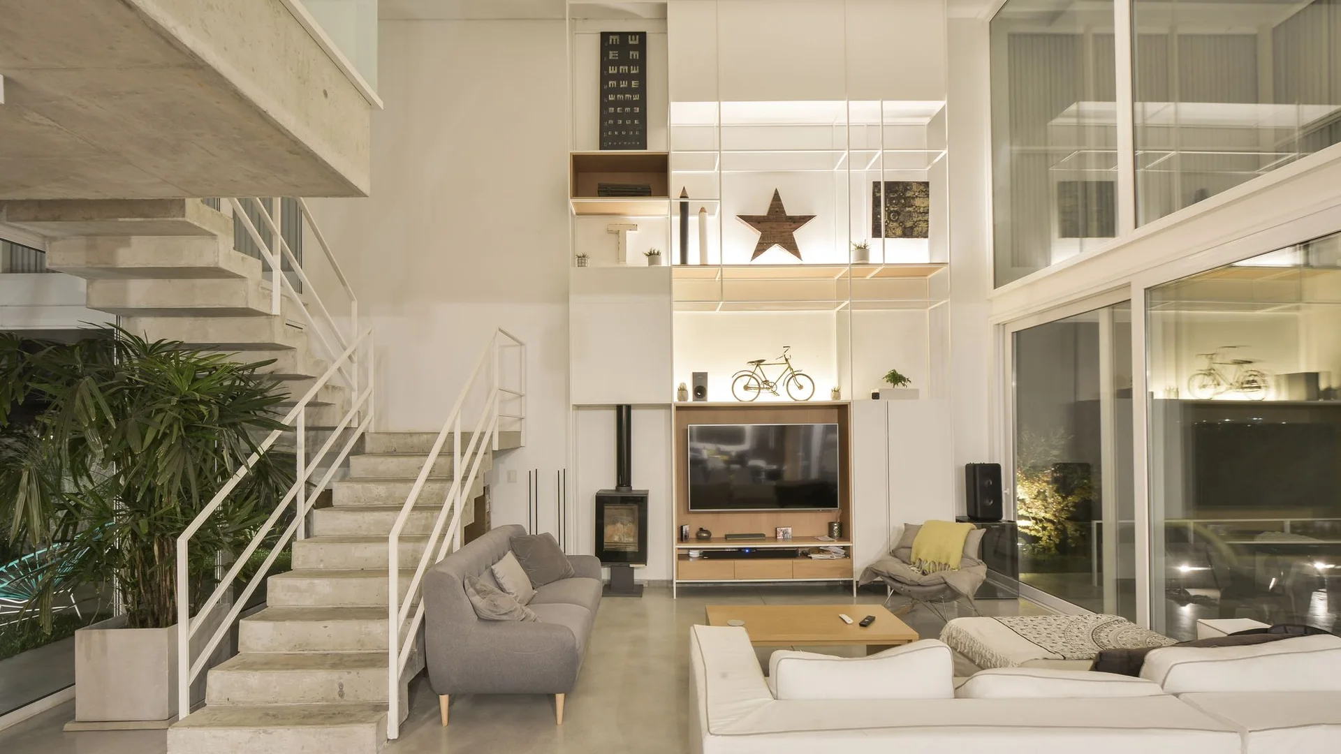 A double-height contemporary living room features a polished concrete floor, a floating concrete staircase, and a tall white metal grid shelving unit.