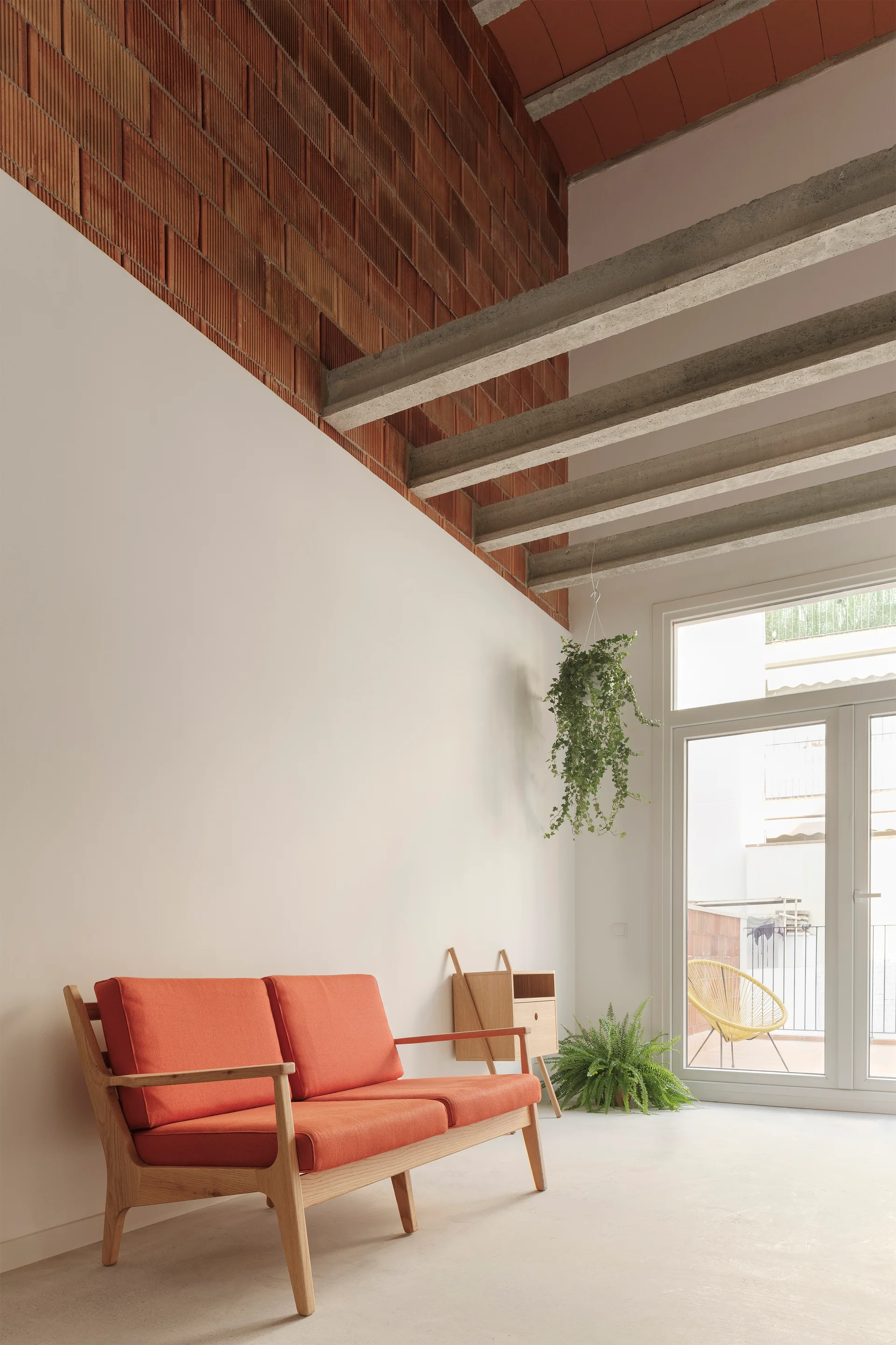 A minimalist living room features a modern orange sofa, white walls, and high ceilings with exposed concrete beams and a red brick wall.