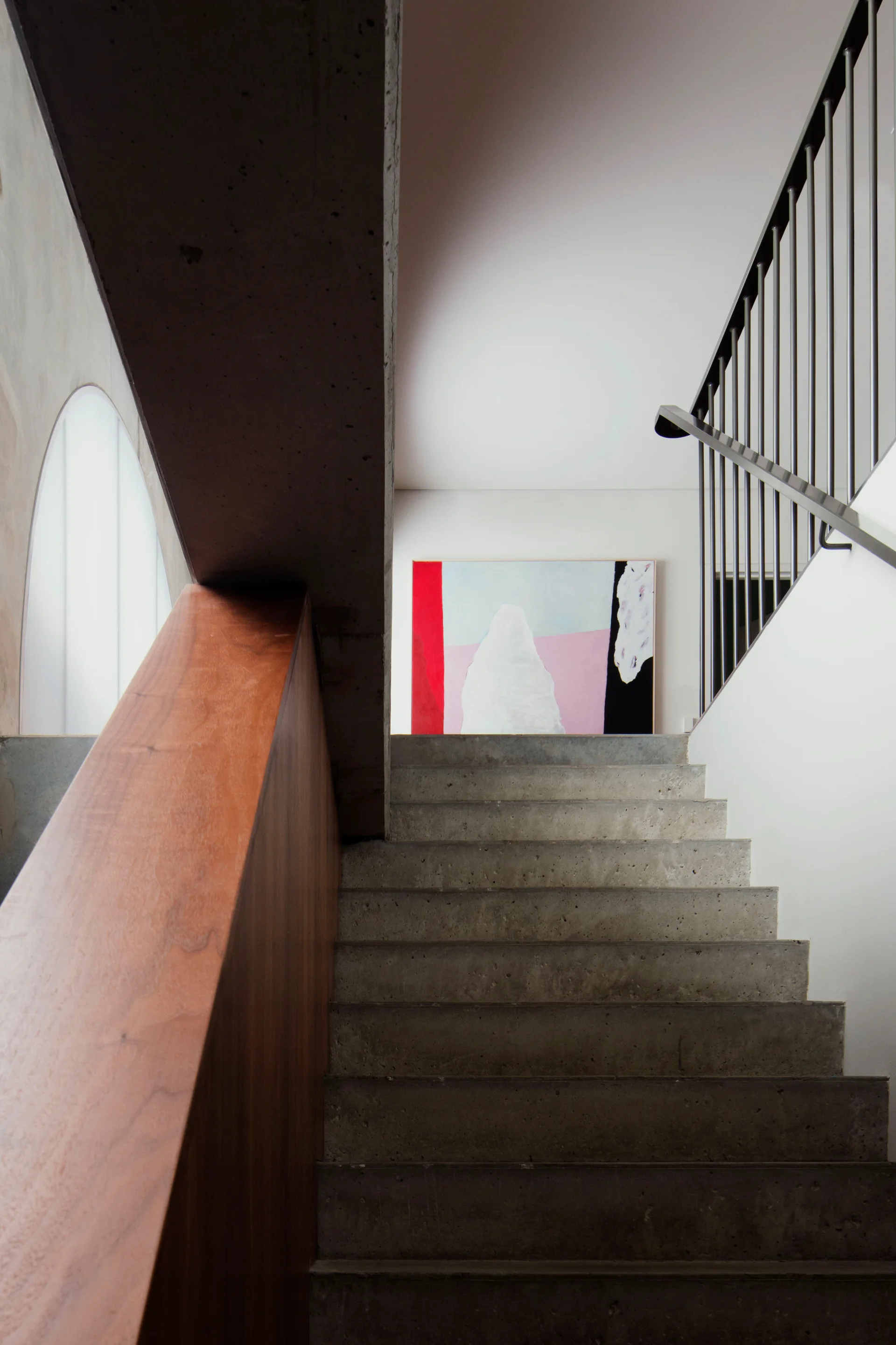 A minimalist concrete staircase with a warm wood balustrade leads up to a large abstract painting on the wall.