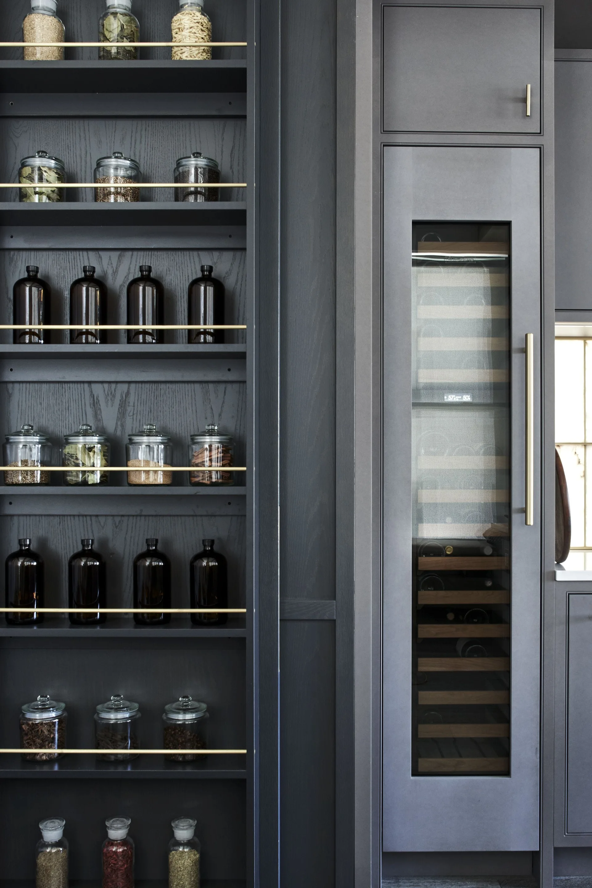 Built-in dark charcoal pantry shelves hold glass jars behind thin brass rails next to an integrated wine refrigerator.