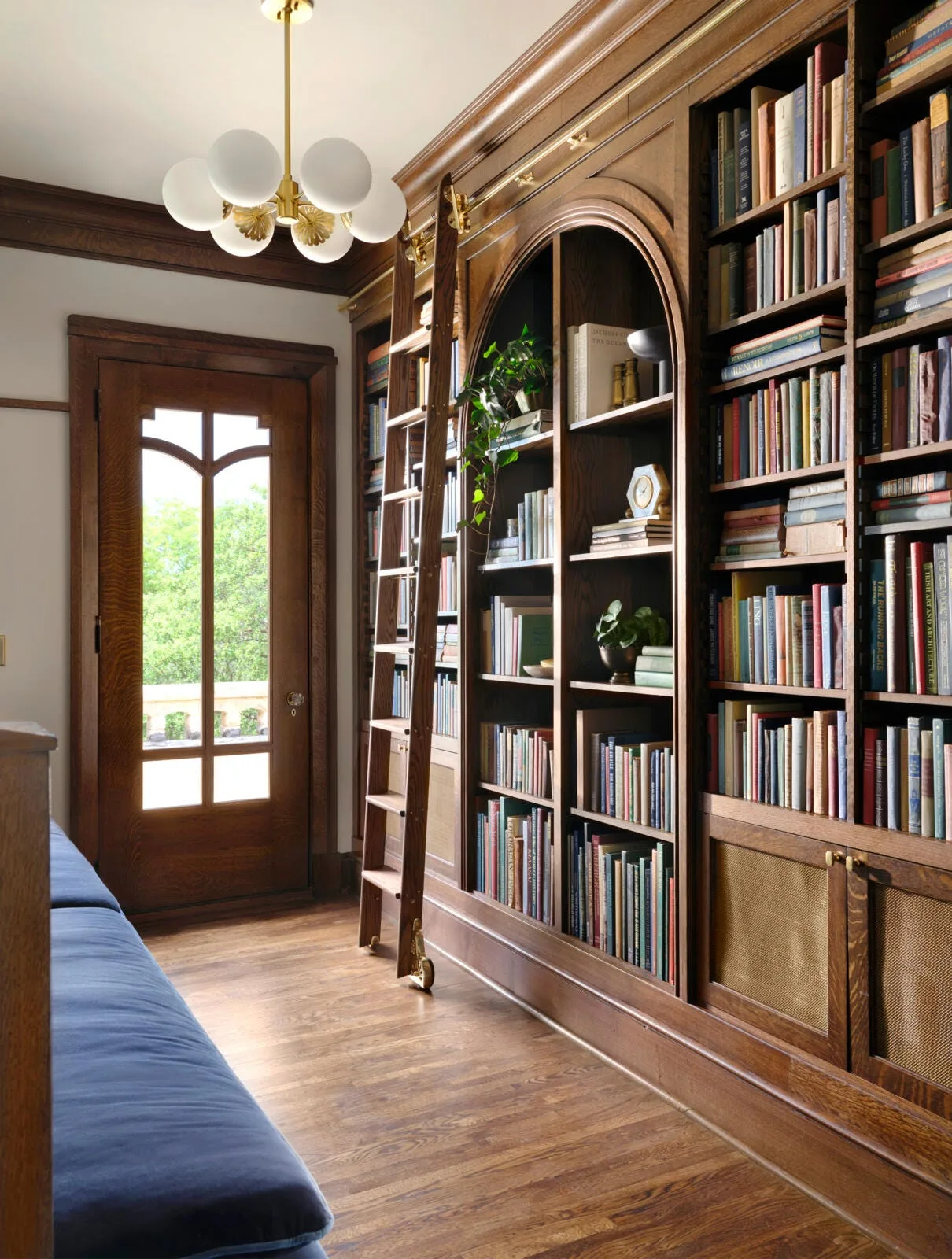 Floor-to-ceiling wooden bookshelves line a home library featuring a rolling wooden ladder on a brass rail and an arched display nook.