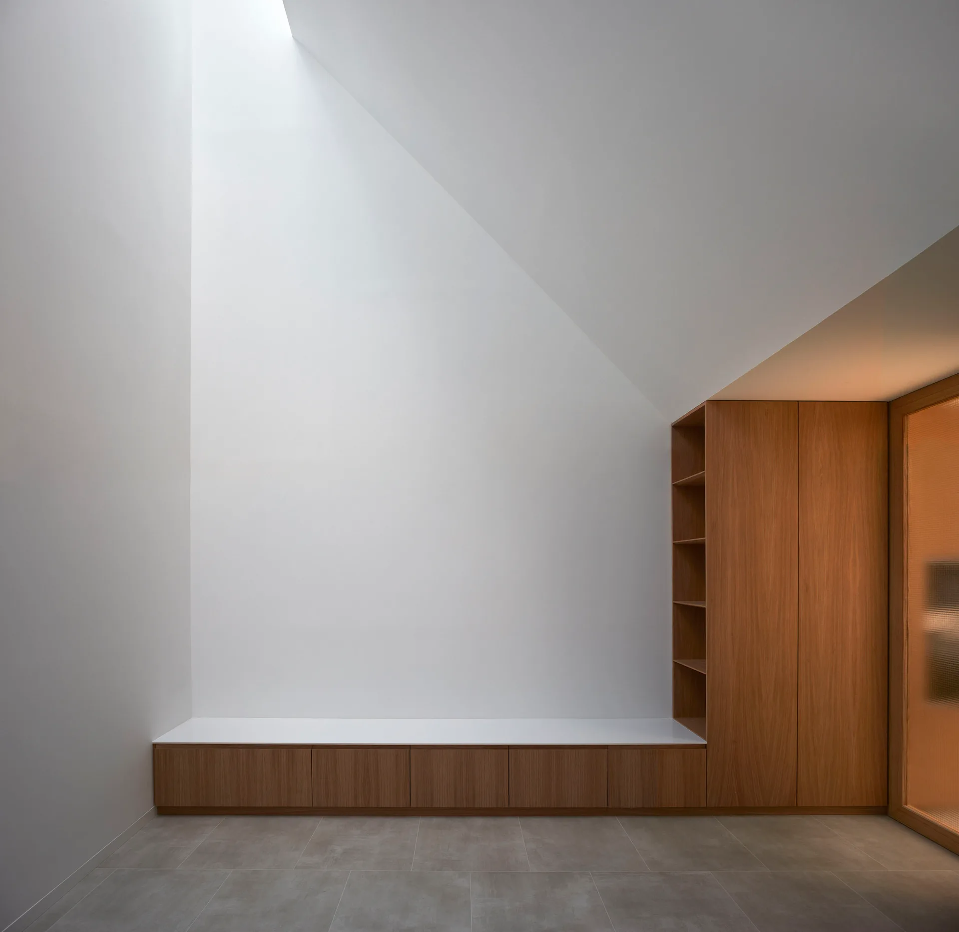 A minimalist room features a long low wooden bench, a tall wood cabinet, and gray tile flooring beneath a large angled skylight casting light.